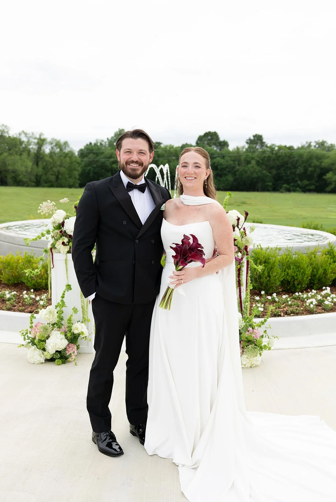  Elizabeth & Ryan smiling at the ceremony setting, fountain and floral pillars behind them 
