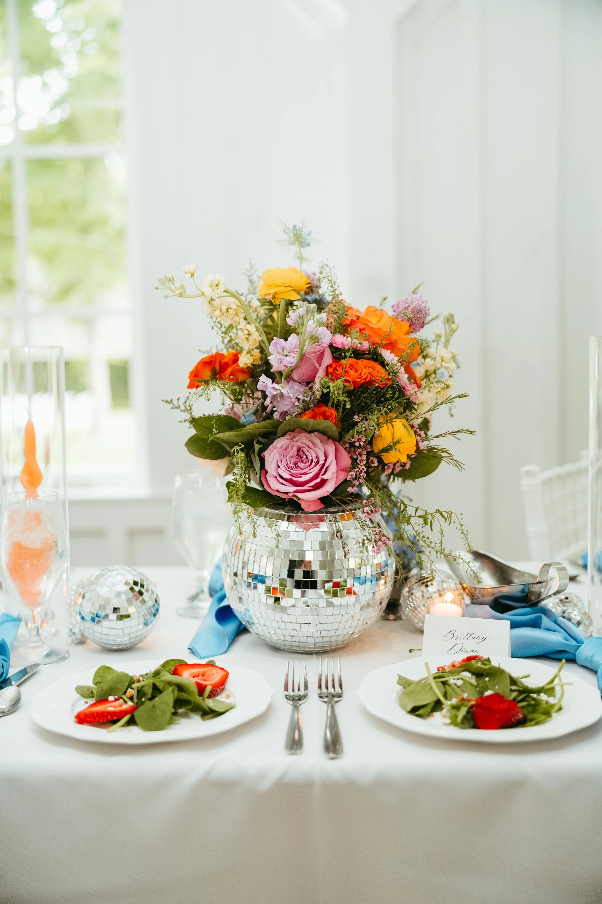 Colorful floral centerpiece in silver vase set on an elegant wedding table with fresh salad plates and blue napkins