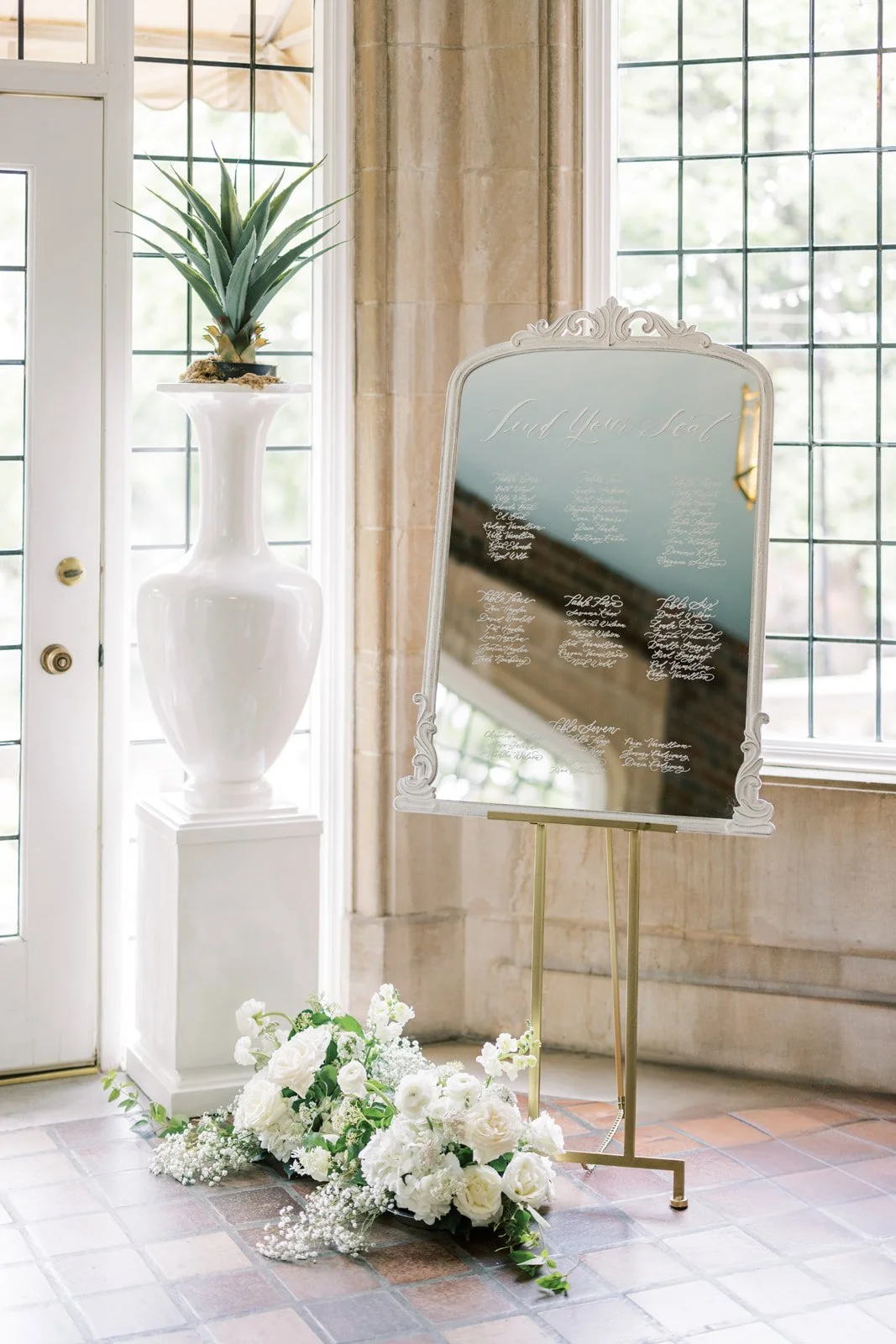 Wedding seating chart displayed on a decorative mirror with floral arrangements at the base and a tall white vase with a plant on a pedestal nearby, in a room with large windows and tiled floor.
