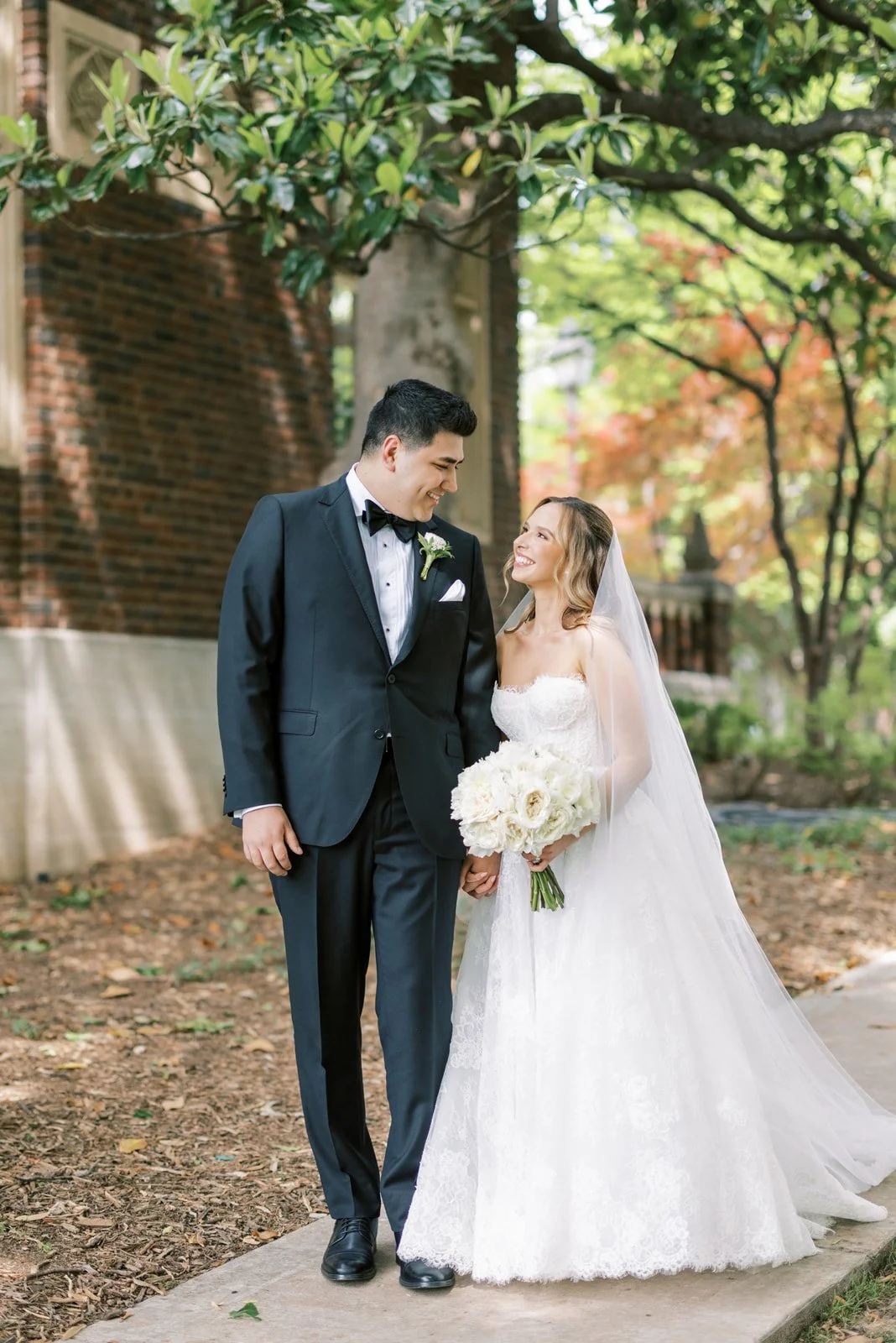 A bride and groom smiling at each other outdoors during daytime, with the bride holding a bouquet of white flowers, surrounded by trees with green and red leaves in the background.