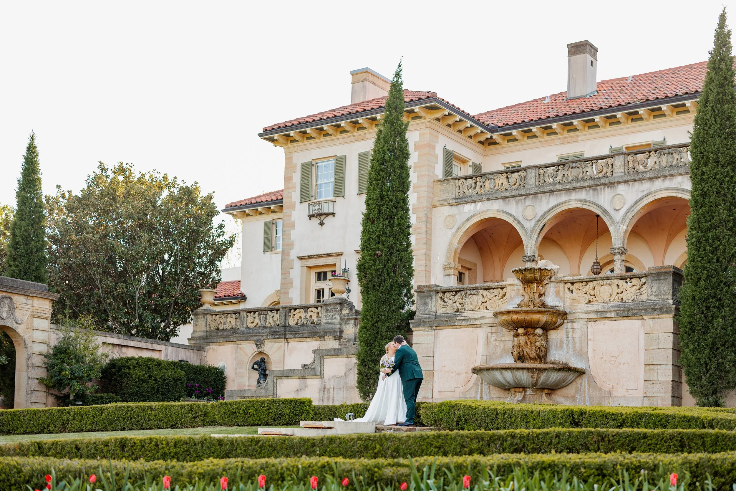 Bride and Groom walk through gardens in front of grand European-style estate with a stone fountain