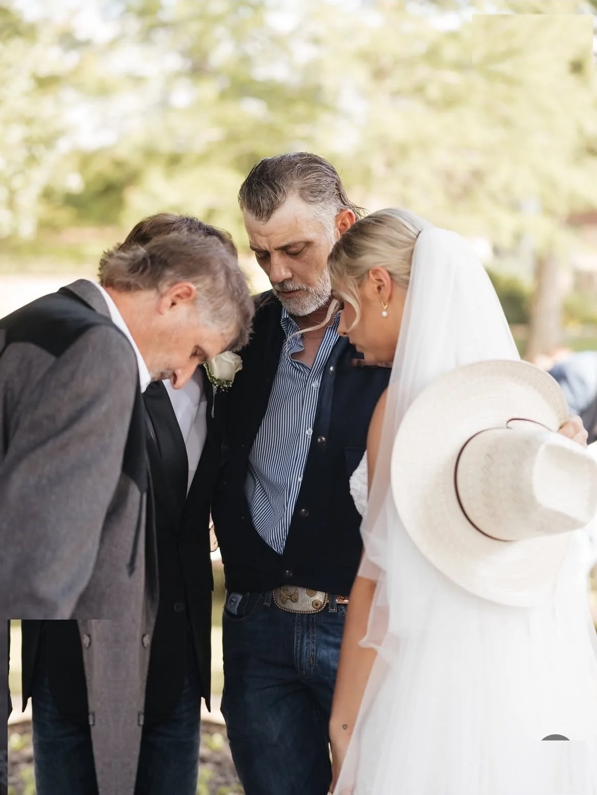  Bride in prayer with family before her wedding ceremony, holding a white cowboy hat 