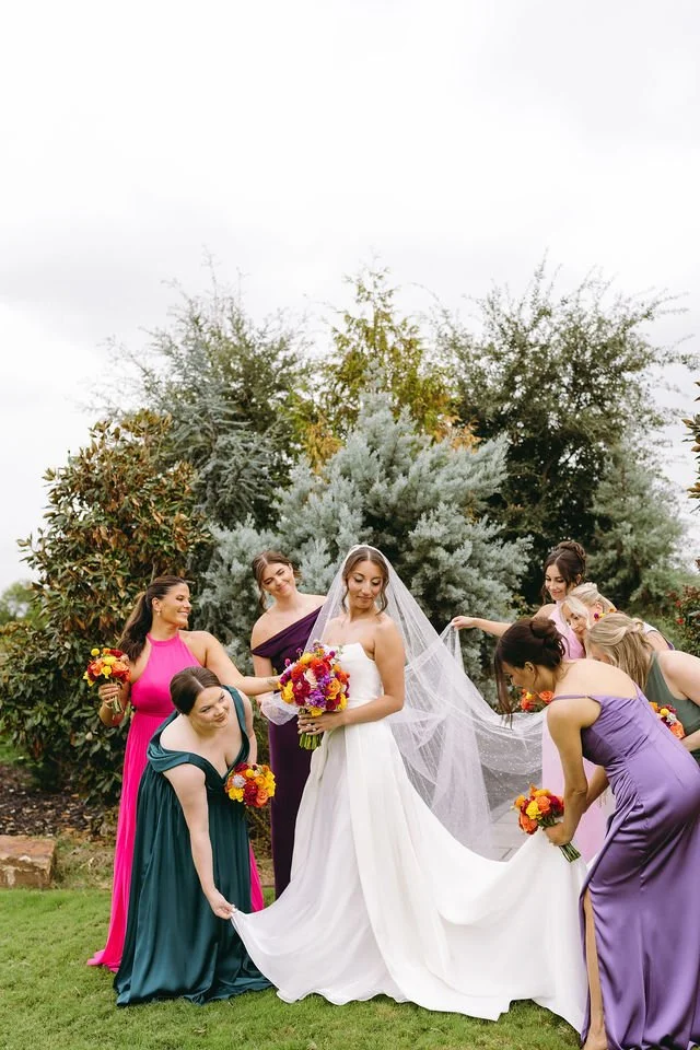 bridesmaids in bright colored dresses helping the bride with her dress and veil
