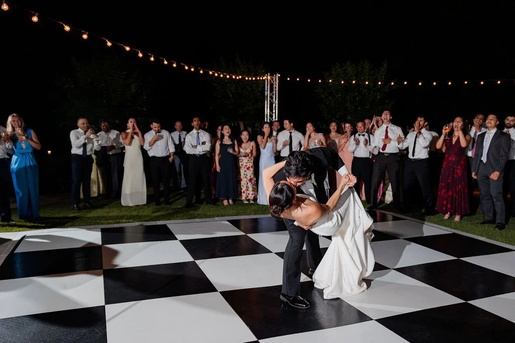 Groom dipping his bride during their first dance on a checkered dance floor under string lights
