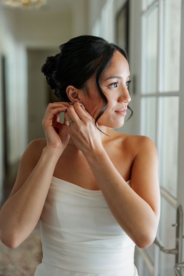 Bride putting on pearl earrings beside a bright window