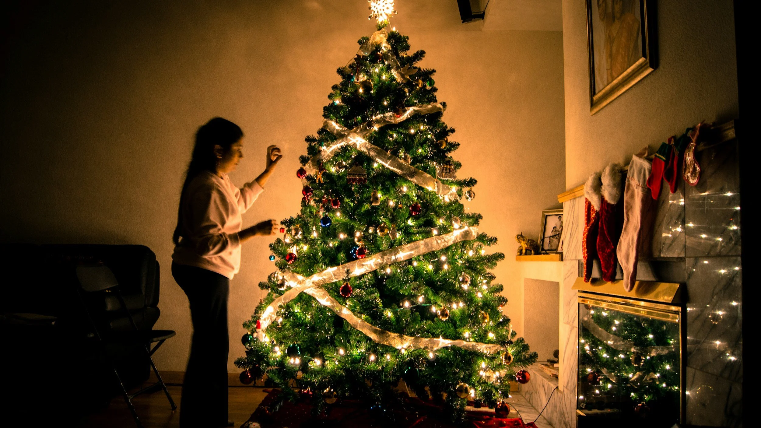 A woman decorating a Christmas tree with lights and ornaments in a warmly lit living room, with stockings hanging on a fireplace mantel and a fireplace reflecting the Christmas tree lights.