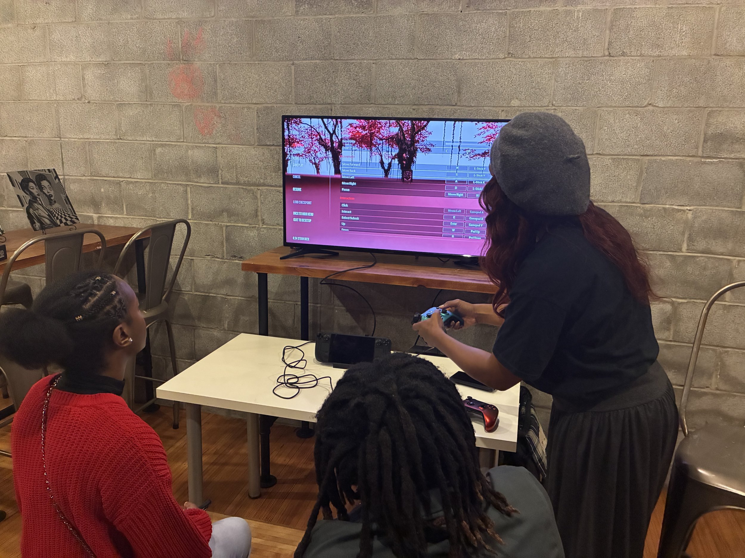 A woman wearing a gray beanie and black shirt is playing a video game with a controller while standing in front of a television displaying a scene with pink trees and a blue sky. She is surrounded by children sitting at a table, with one wearing a red sweater in the foreground and another with braided hair.