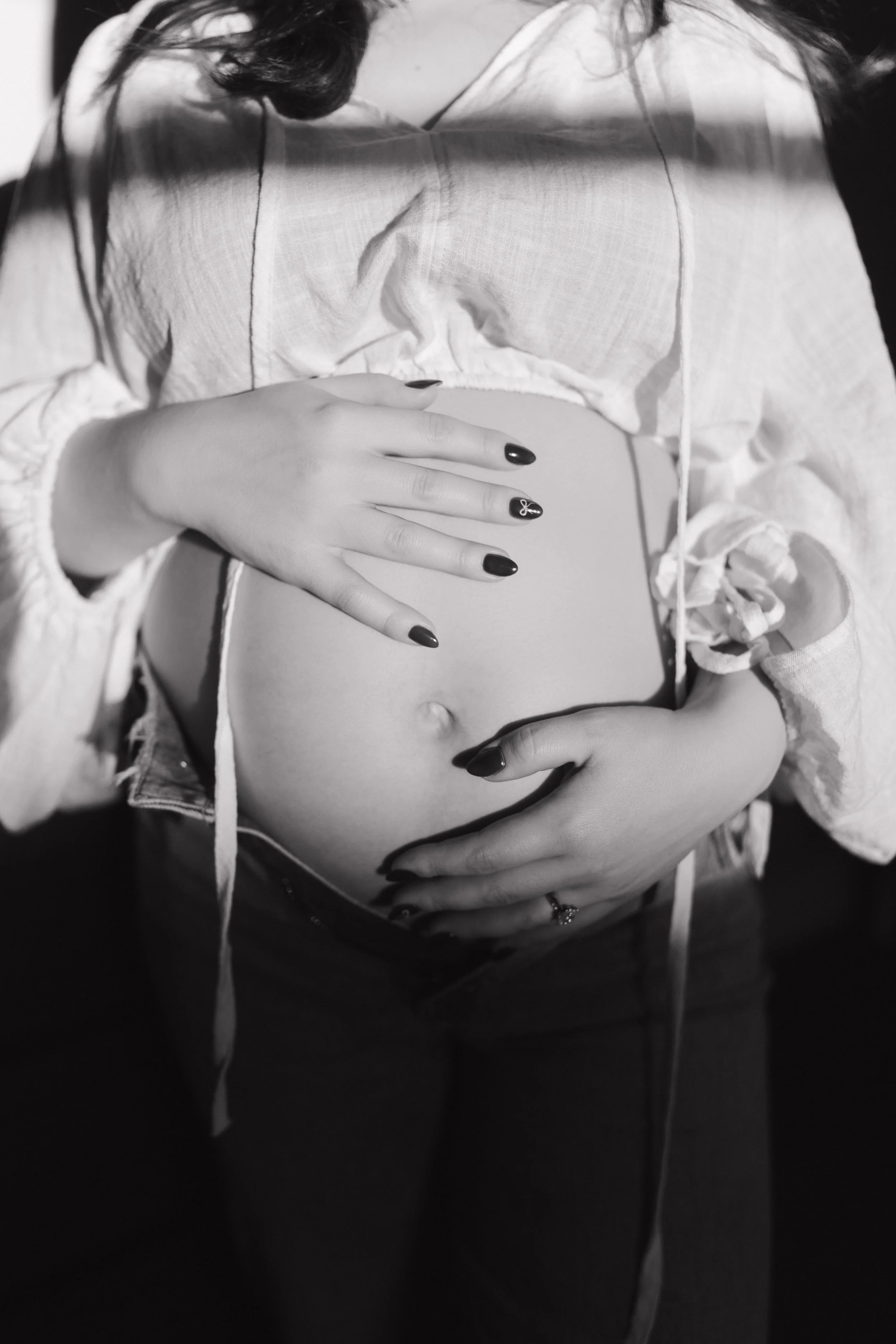 A woman with dark hair, wearing a light-colored, long-sleeve, textured top, is holding her bare pregnant belly with both hands. Her nails are painted dark, and she is wearing rings on her fingers. The image is black and white.