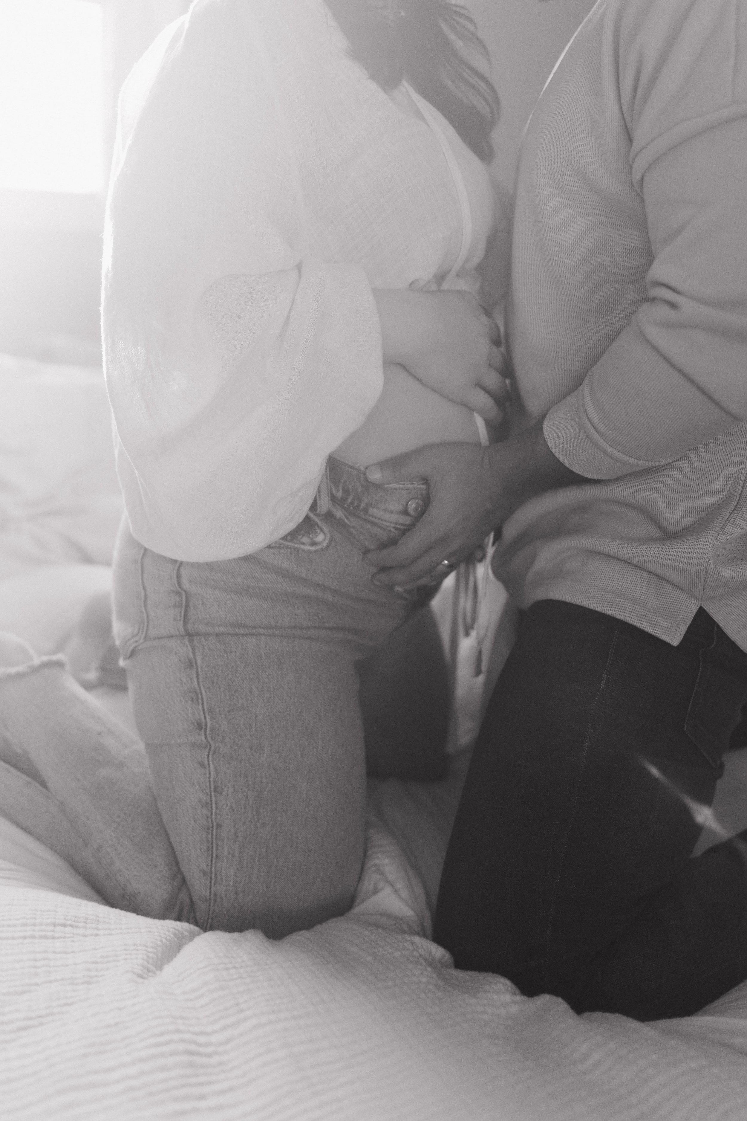 A pregnant woman kneeling on a bed, with a man holding her belly and kissing her stomach.