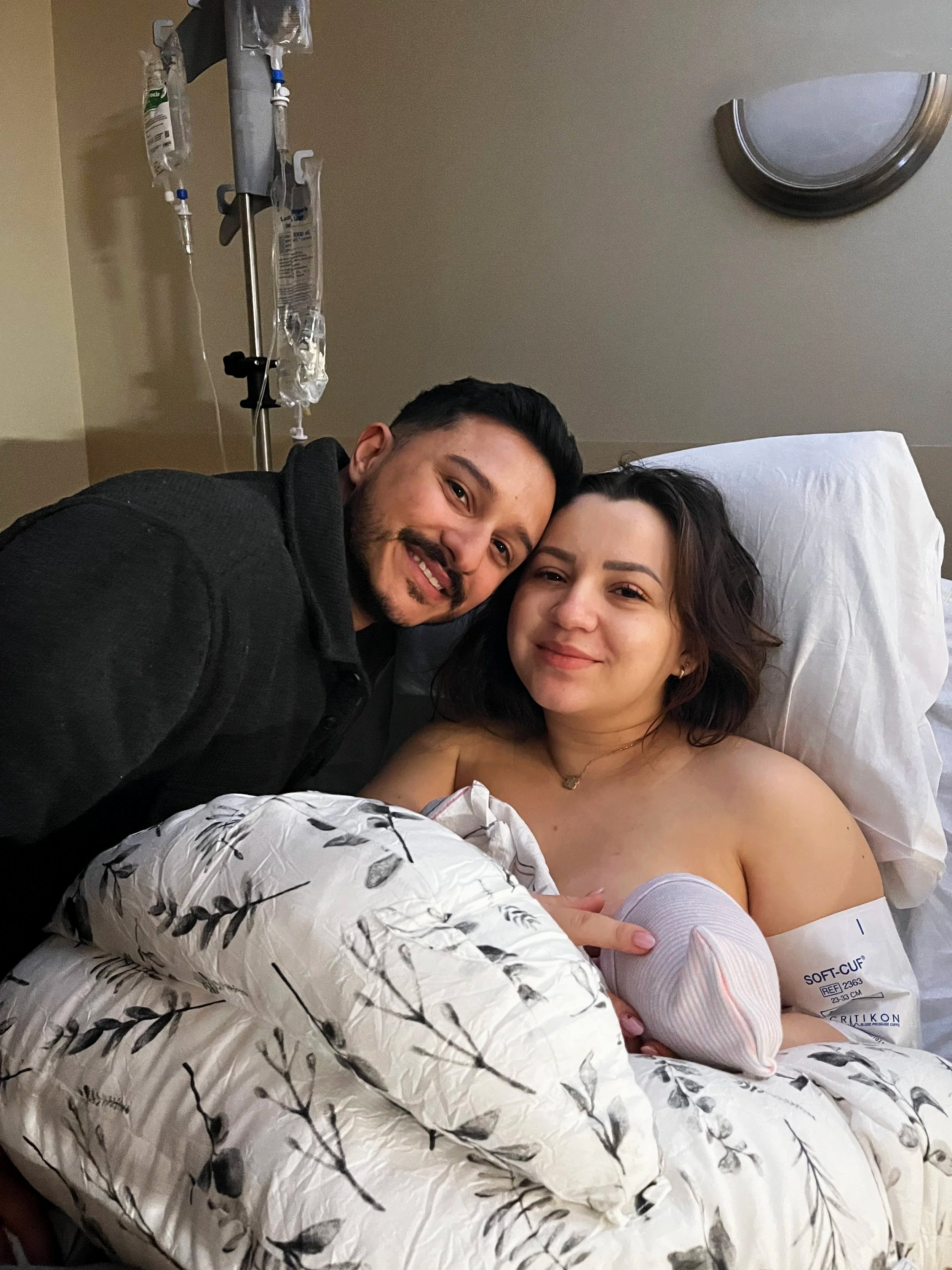A woman lying in a hospital bed, holding her newborn baby, with a man leaning in beside her, all smiling. Medical equipment is visible in the background.