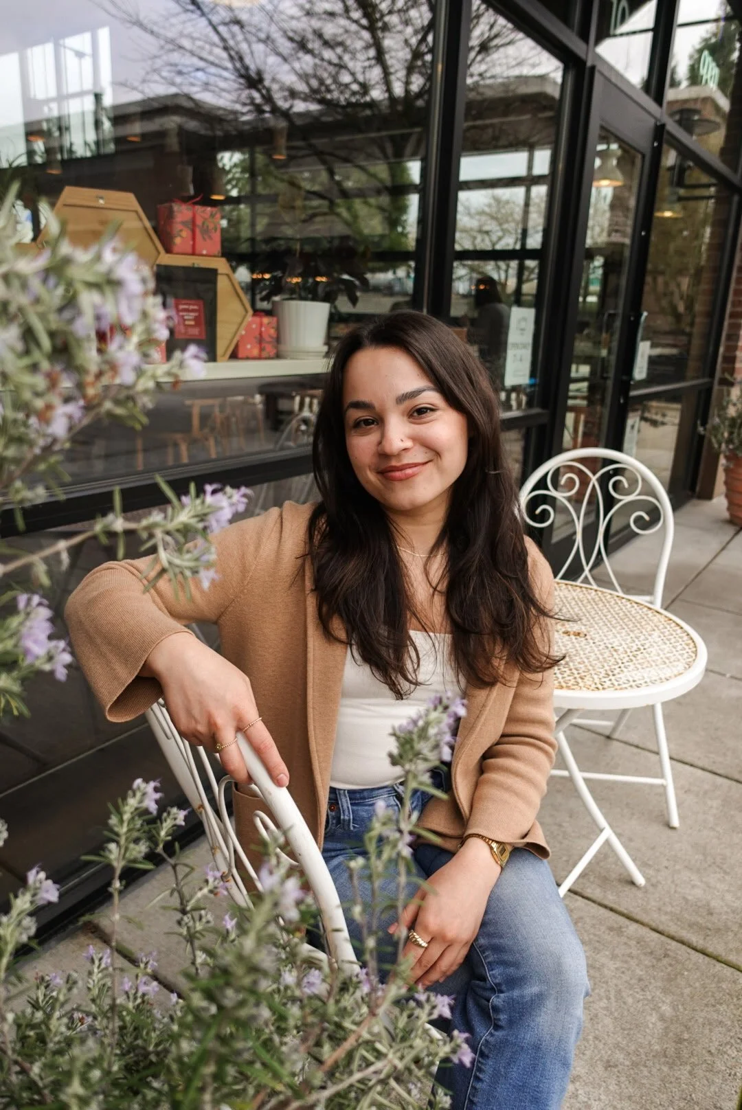 A woman with long dark hair and light skin sitting outdoors at a white metal chair, smiling at the camera, with a box and plants in the background.