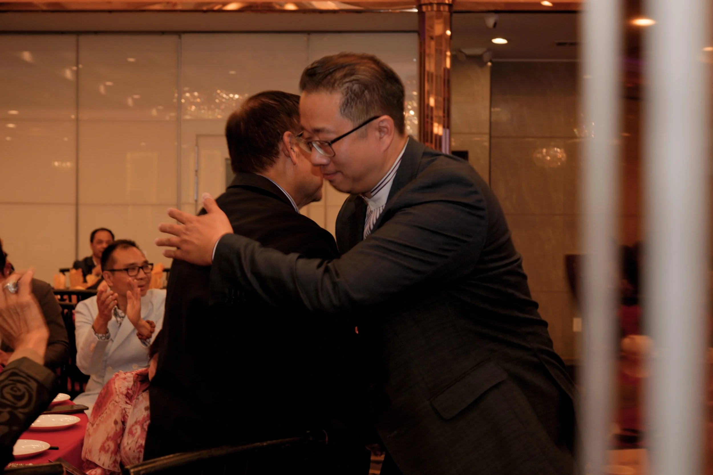 Two men hugging at a formal event, with seated audience members clapping and smiling in the background.