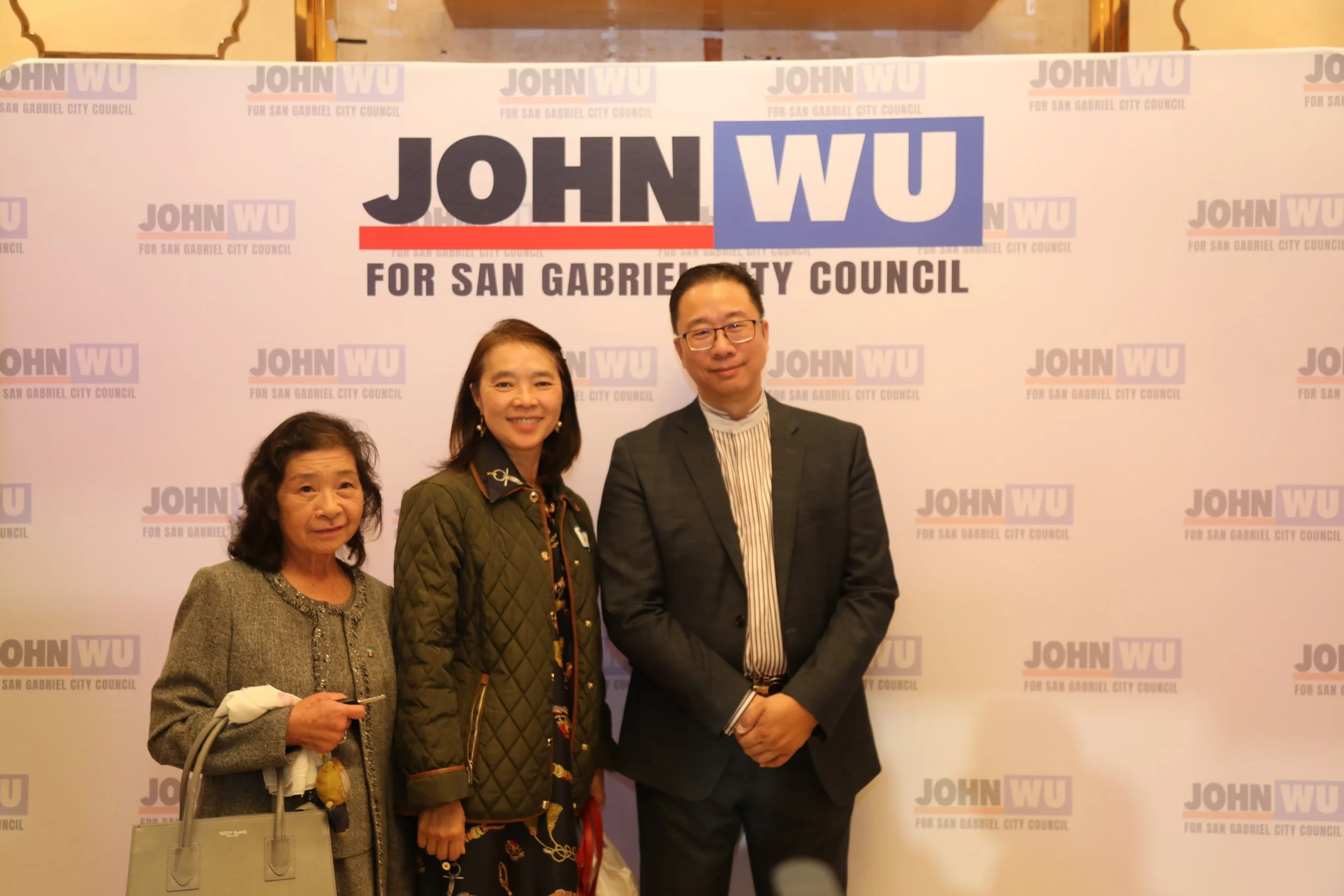 Three individuals at a political event standing in front of a backdrop that reads 'JOHN WU for San Gabriel City Council.' The individuals are smiling; the woman on the left is holding keys and a purse, the woman in the middle is wearing a green quilt
