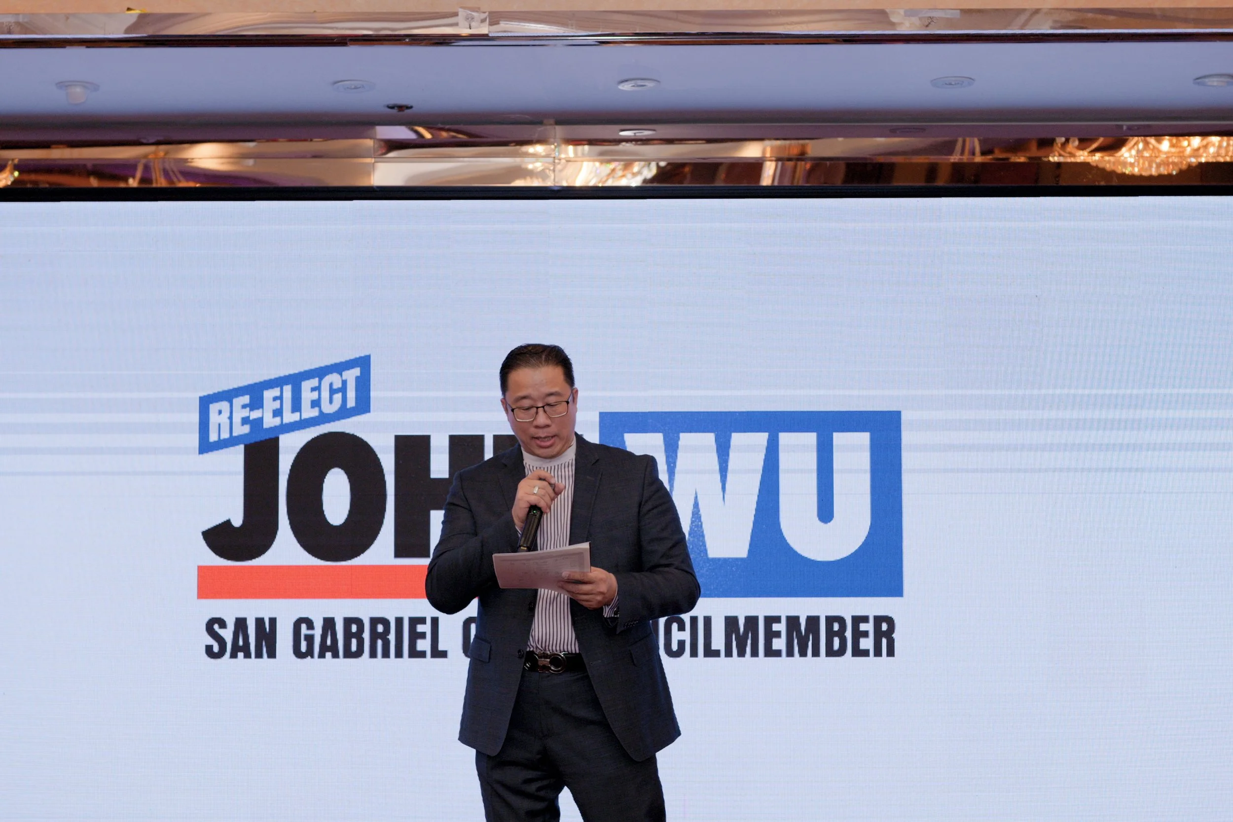 Man in a dark suit and glasses speaking into a microphone in front of a large screen with a political campaign poster reading 'Re-Elect Joe J. V.' and 'San Gabriel'.