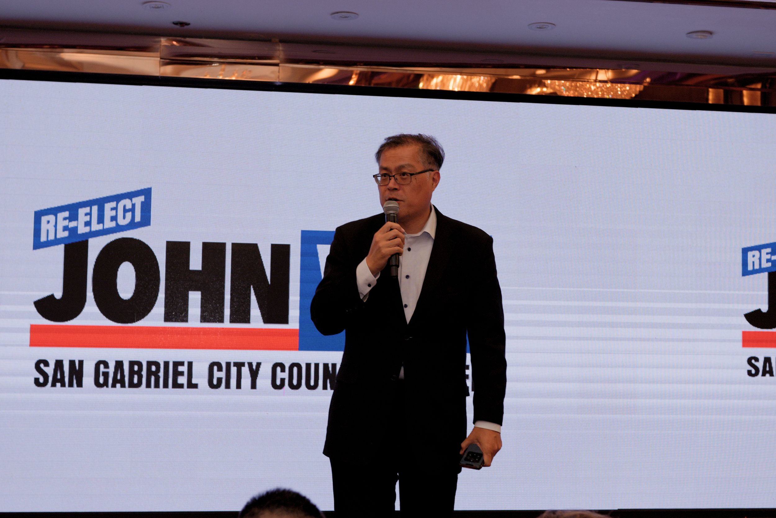 A man in a black suit and glasses standing on stage holding a microphone, speaking at an event about San Gabriel City Council, with a large screen behind him displaying the words 'RE-ELECT' and 'JOHN'.