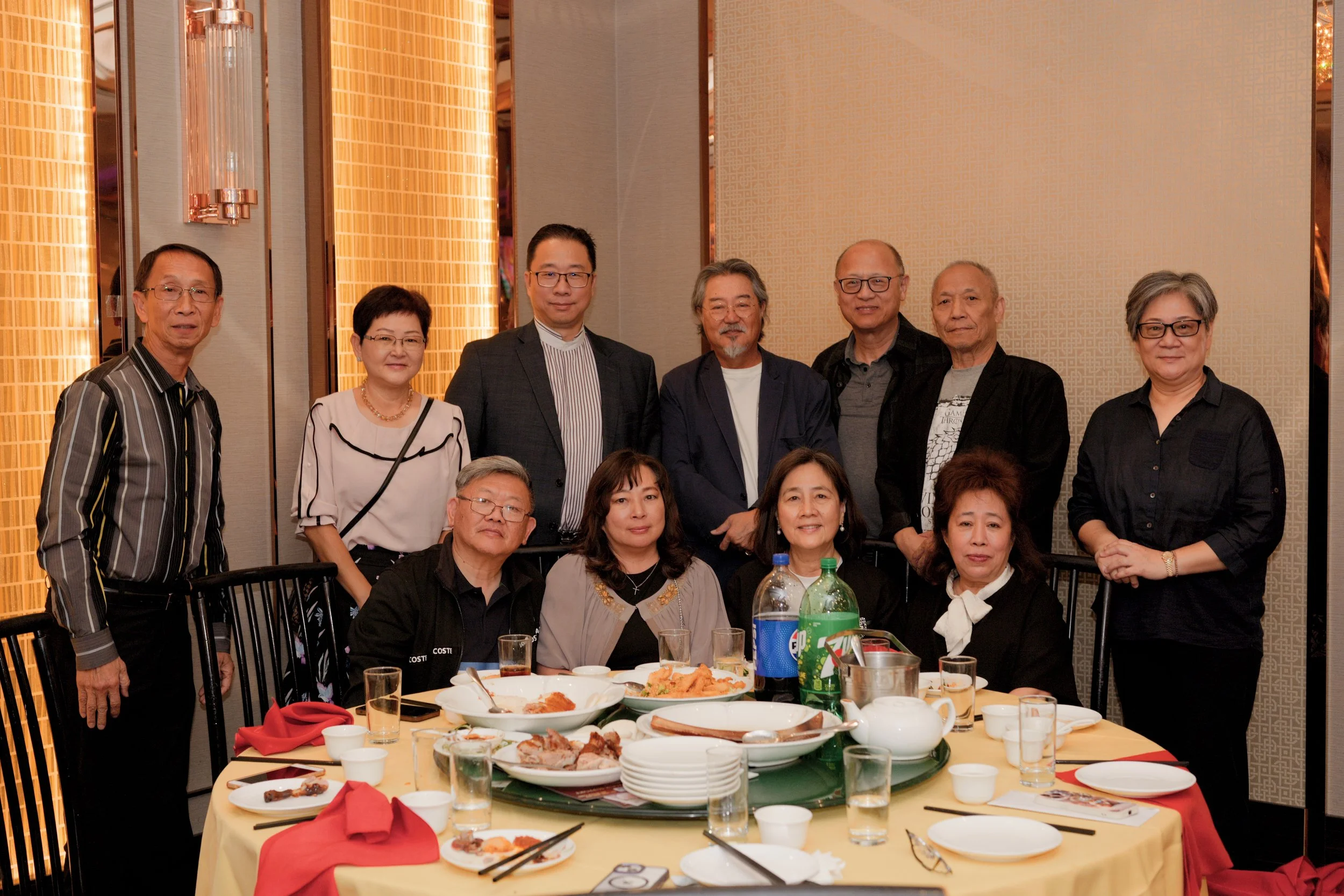 A group of people gathered around a dining table at a celebration or event, with food and drinks on the table, inside a warmly lit room.