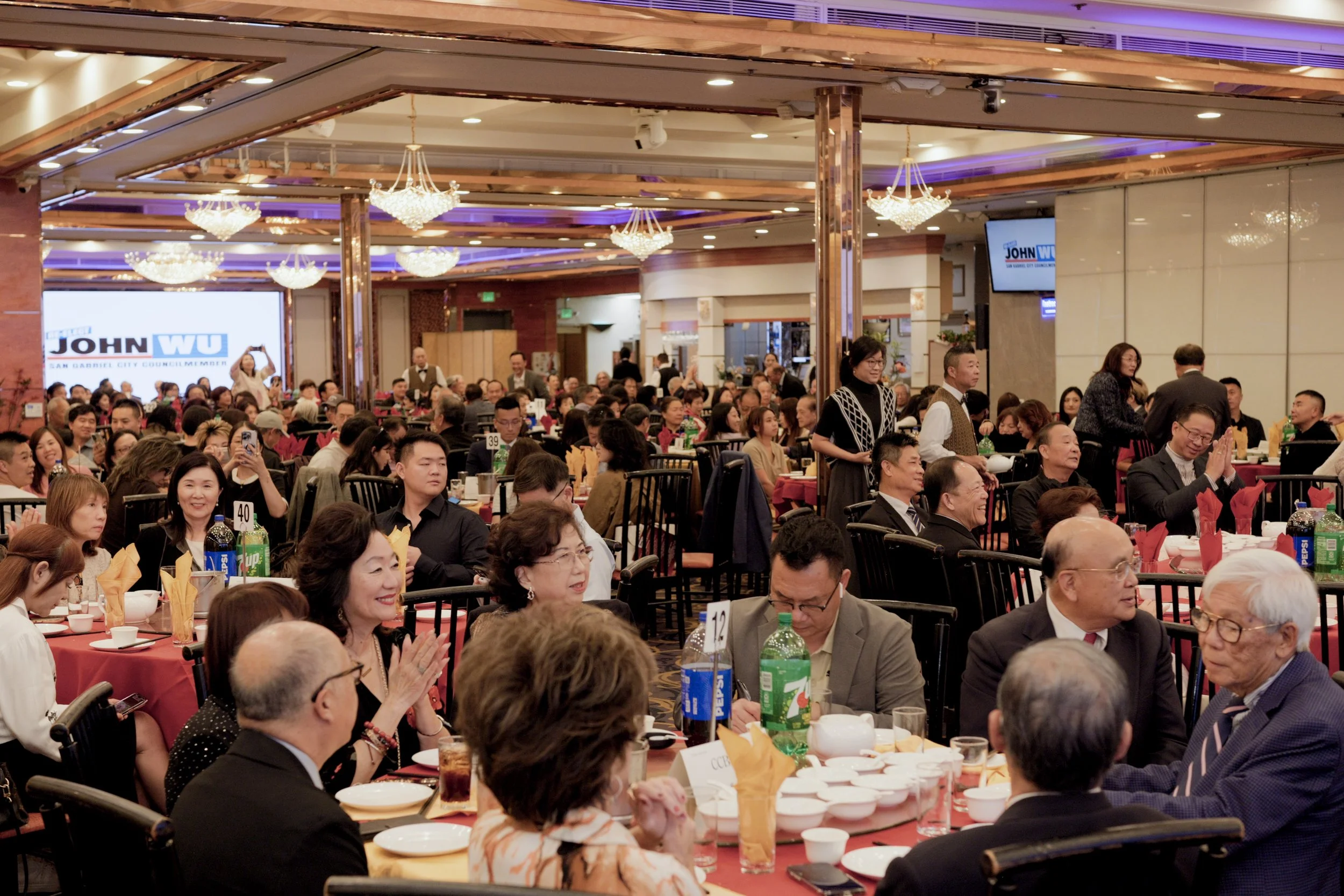 A large indoor event with many people seated at round tables with red tablecloths, eating and talking. The room has chandeliers, purple lighting accents, and two large screens displaying the logo 'JOHN WU, San Gabriel City Council Member.' Attendees 