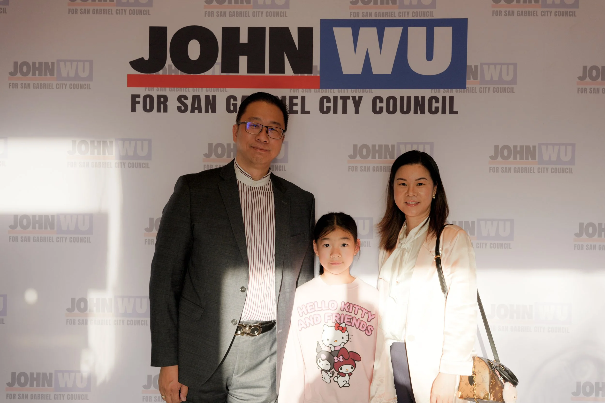 A group of four people, including two adults and two children, standing together in front of a campaign backdrop that reads 'JOHN WU FOR SAN GABRIEL CITY COUNCIL'.