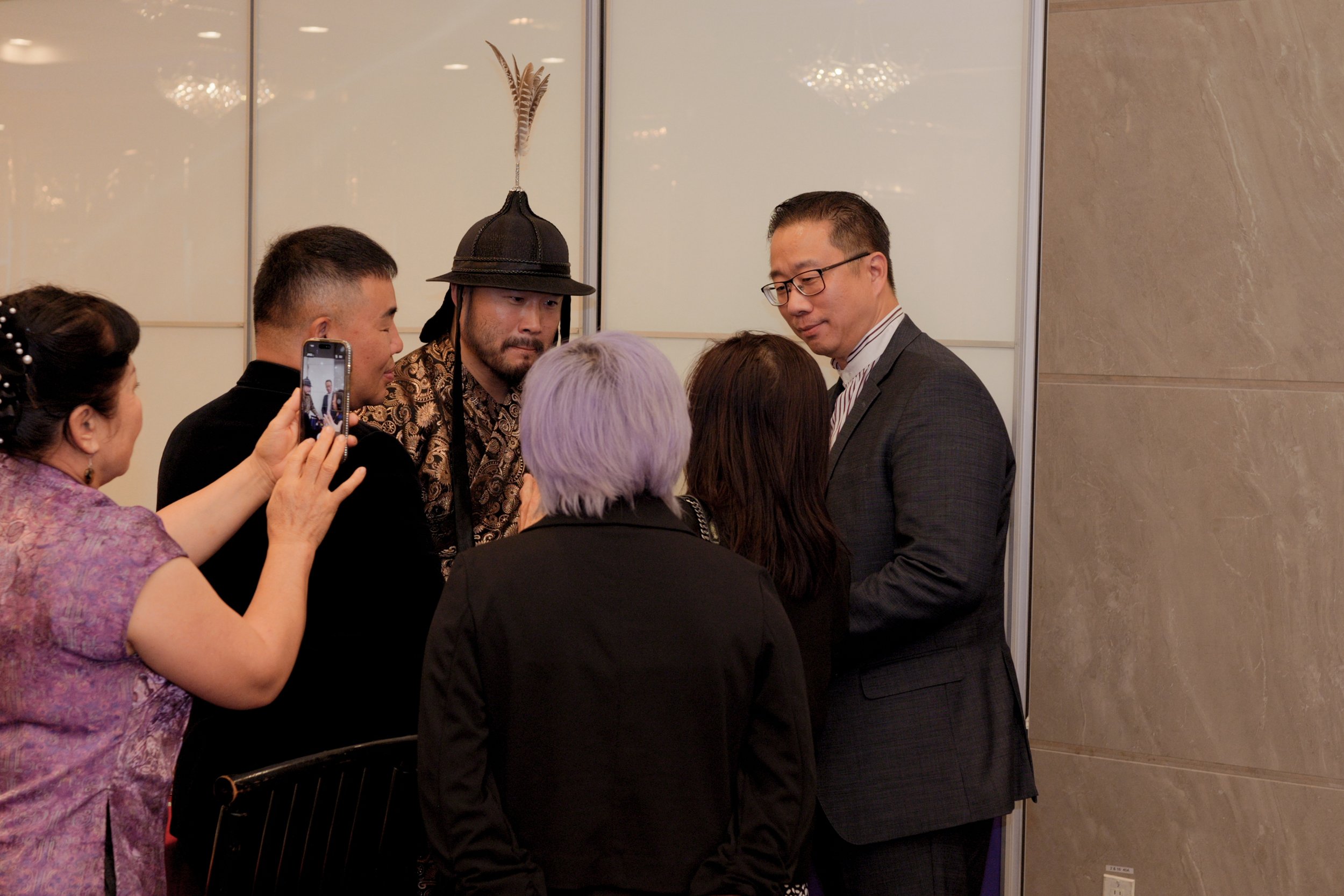 A group of people, including men and women, gather around a man in a suit at an indoor event. One woman is taking a photo with her smartphone. The background features a light-colored wall and a marble column.