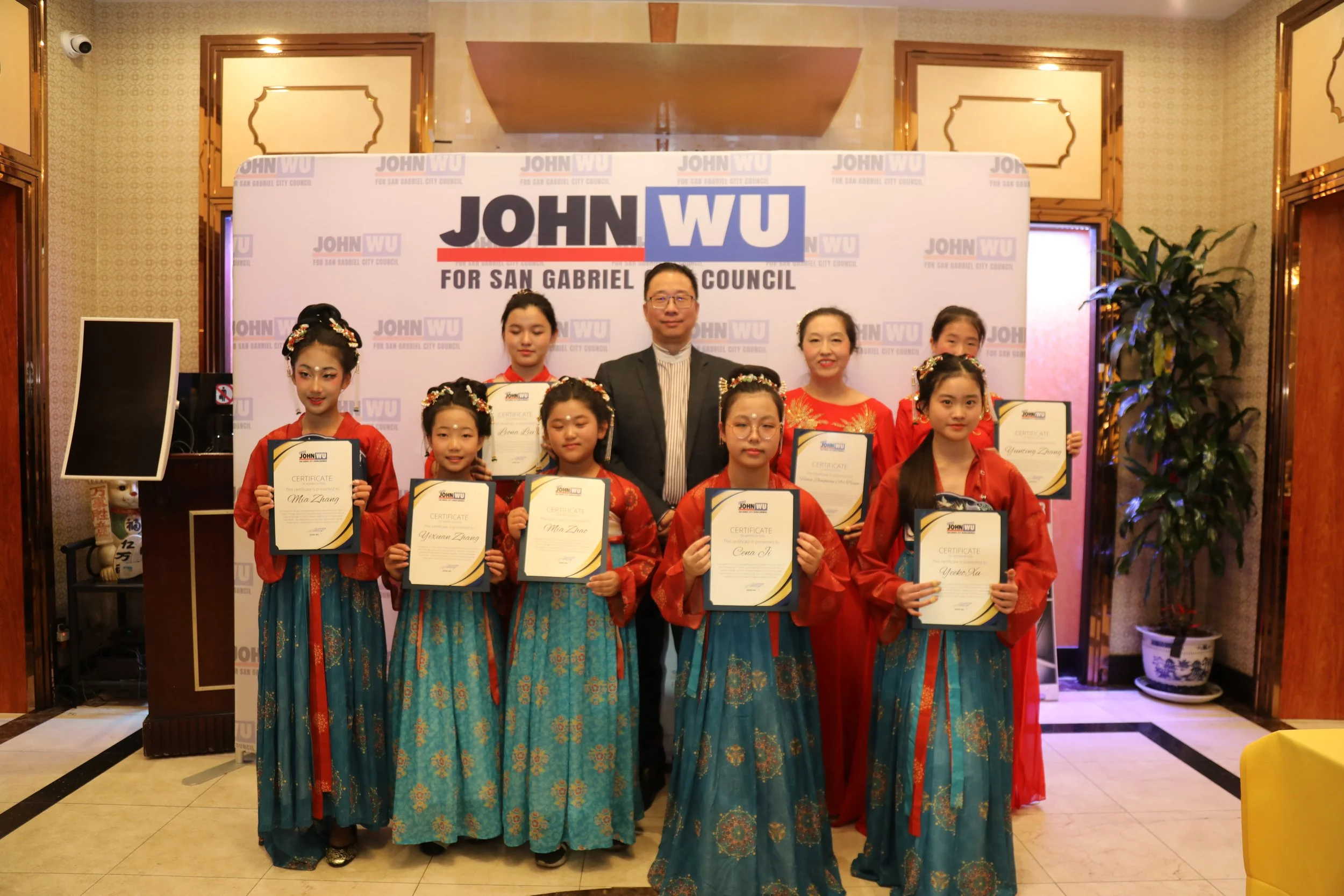 Group of young girls in traditional dresses holding certificates, flanked by a man and a woman, at an award ceremony for San Gabriel City Council, with a backdrop displaying the names 'John Wu' and 'San Gabriel City Council'.