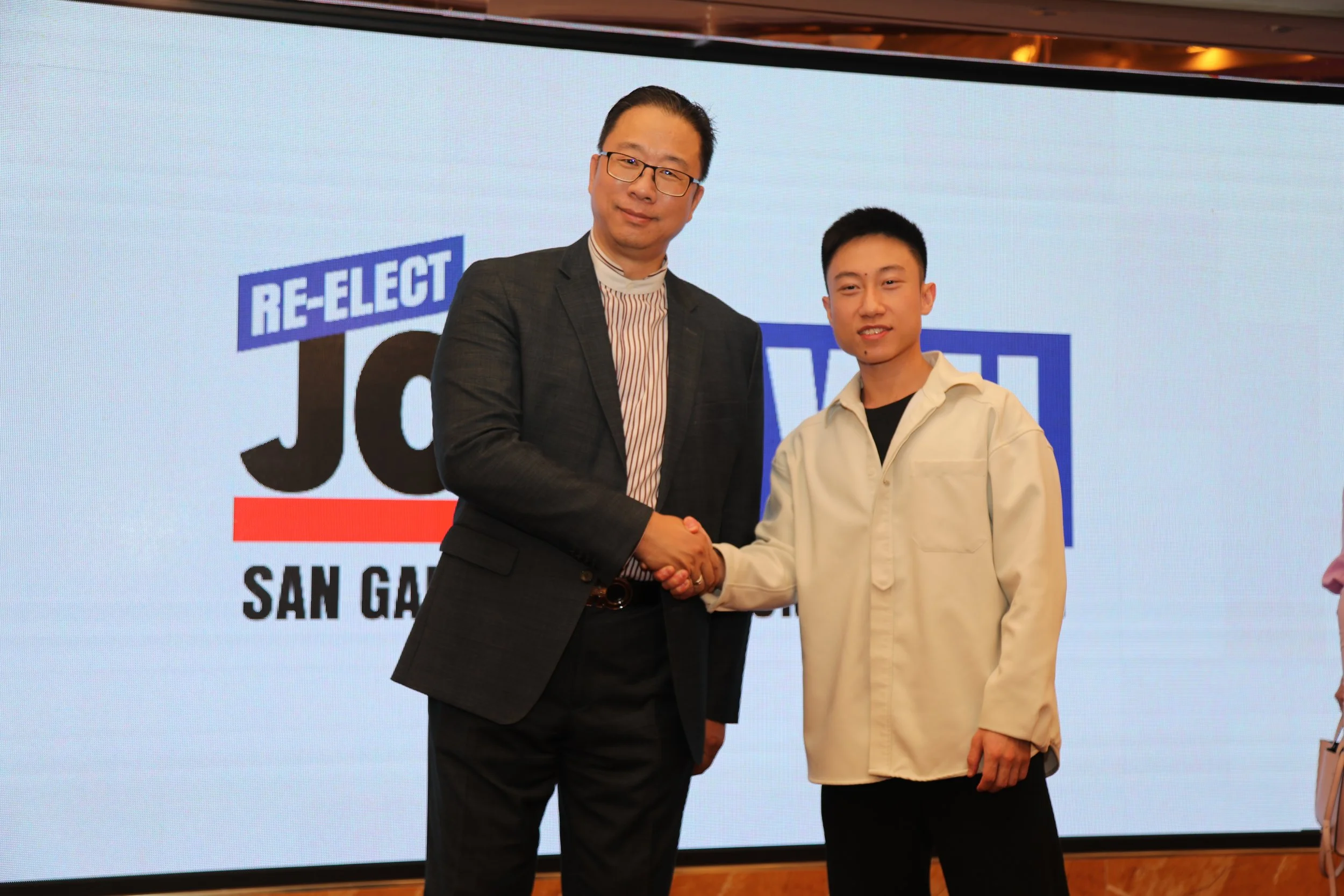 A man in a checked blazer shaking hands with a young man in a beige jacket in front of a large screen displaying a logo that says "RE-ELECT". The background also has text indicating the event location, San Gabriel.