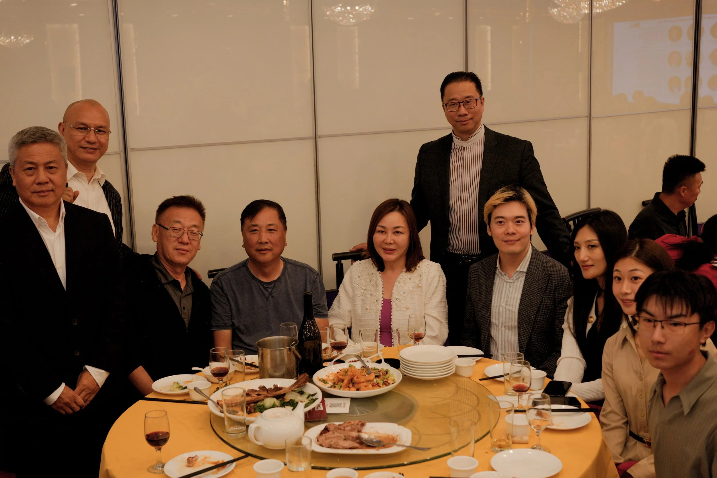 A group of people gathered around a dining table at a restaurant or banquet setting with food and drinks, some smiling and looking at the camera, with a plain wall background.