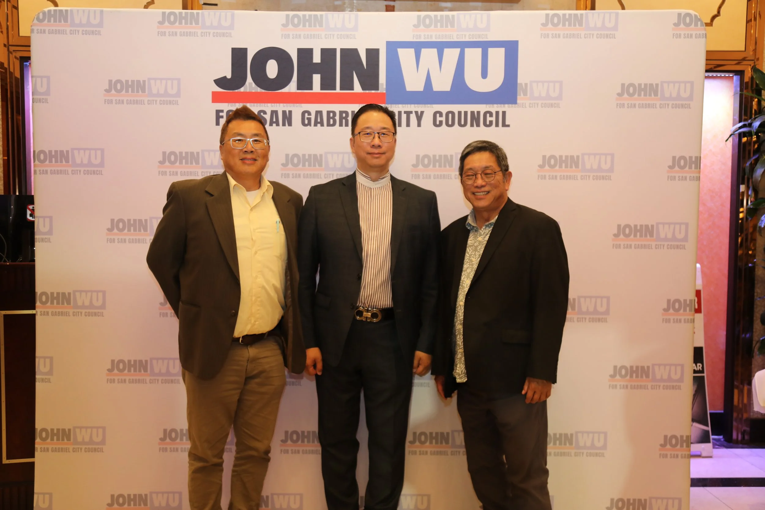 Three men standing together and smiling in front of a backdrop that reads 'John Wu for San Gabriel City Council.'