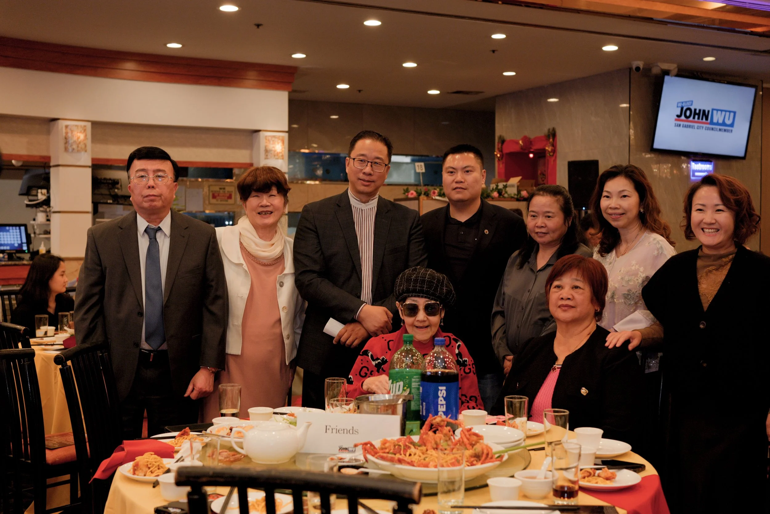 A group of seven diverse people, including men and women, gathered at a dinner event in a banquet hall. They are standing behind a table filled with various dishes and drinks, smiling and posing for the camera. The background shows a large TV screen 