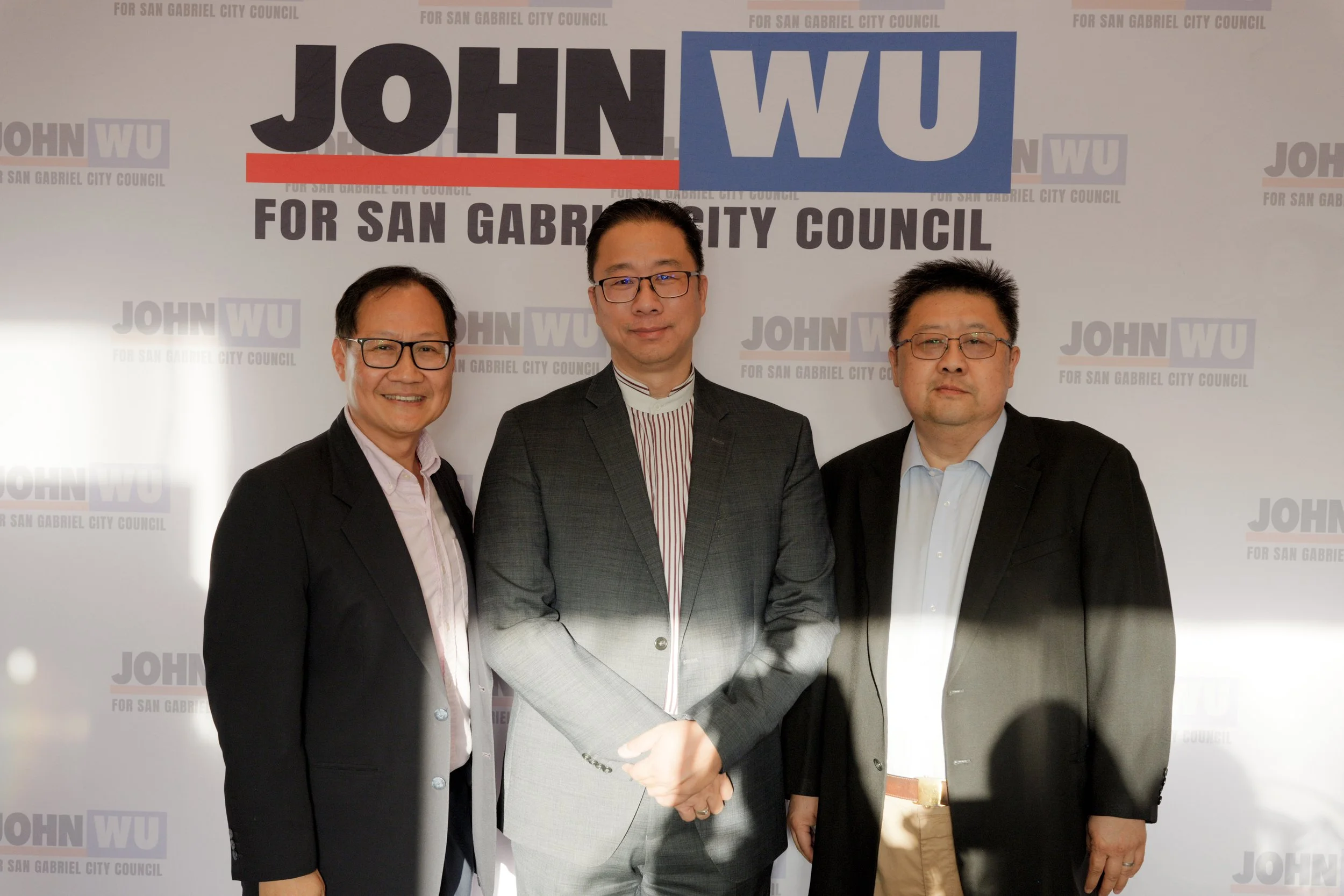 Three men standing together and shaking hands in front of a backdrop that says "JOHN WU FOR SAN GABRIEL CITY COUNCIL" in large letters. All three are dressed in formal attire and wearing glasses.