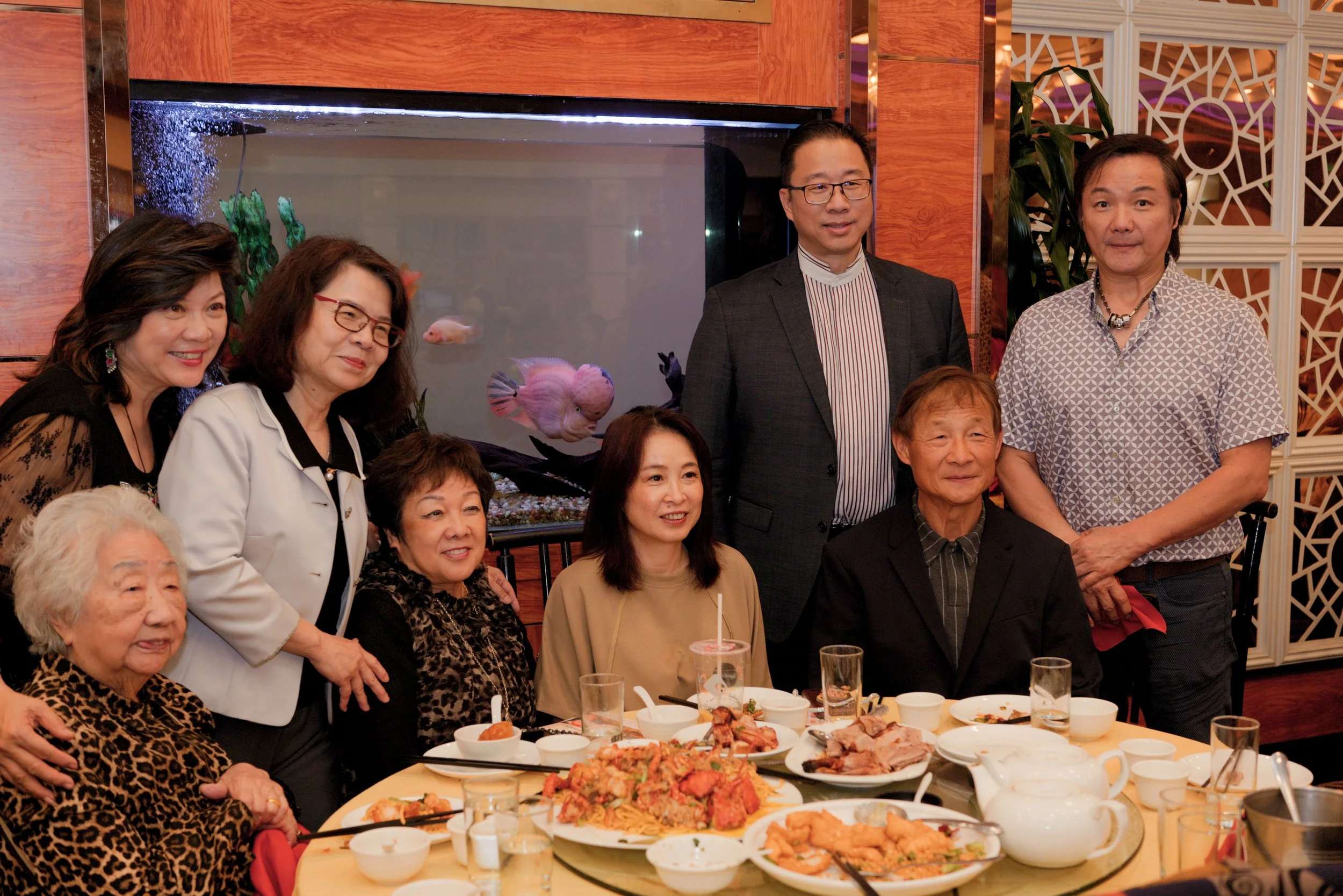 A group of Asian people gathered around a dinner table at a social event, with dishes of food on the table and a large fish tank in the background.