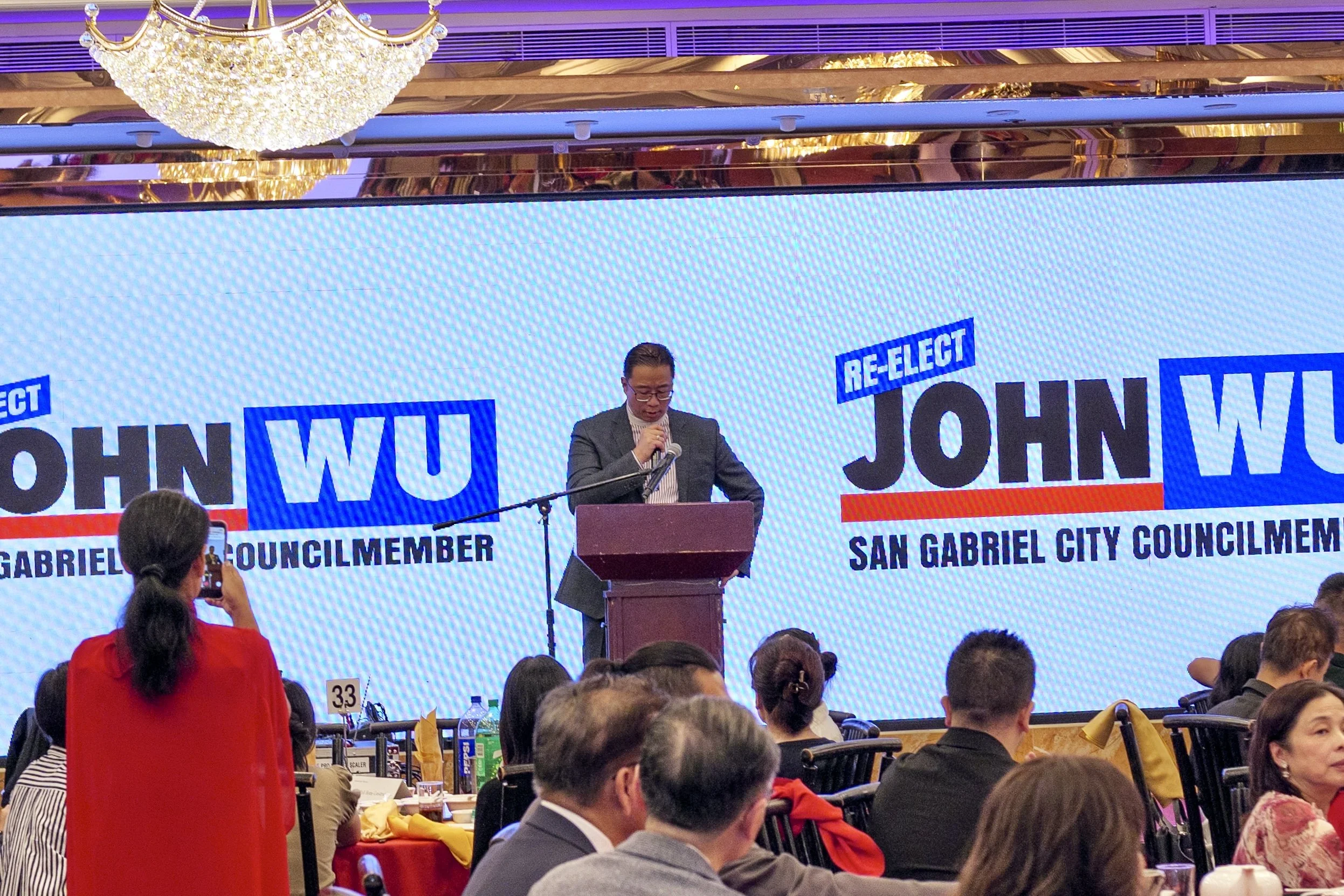 A man standing at a podium giving a speech during an event. The large screen behind him displays text about re-election and his name, John Wu. There is an audience seated and some appear to be taking photos.