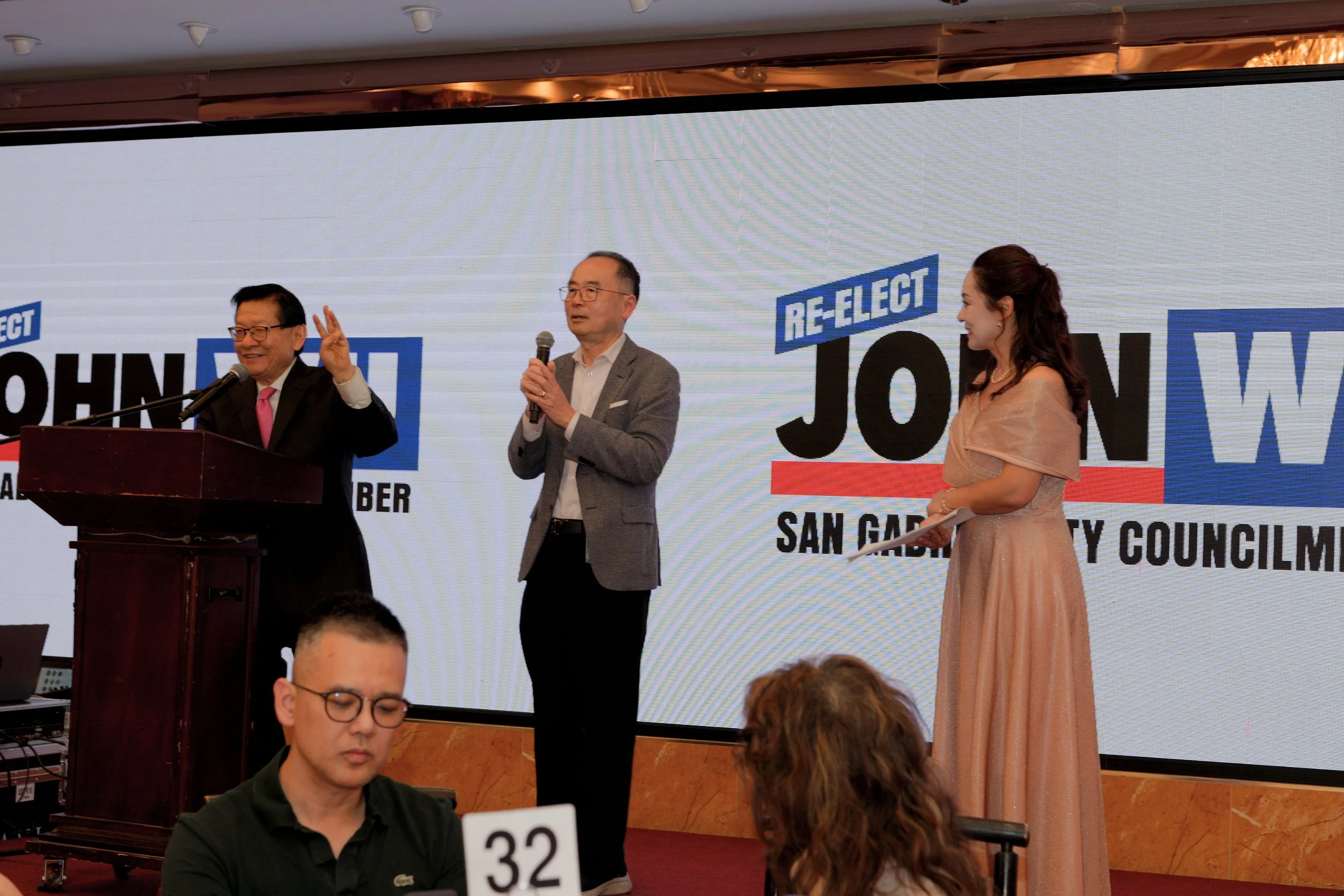 A group of three people standing in front of a large screen with the words 'RE-ELECT JOHN DOE SAN GABRIEL CITY COUNCIL' visible. Two men are holding microphones, while a woman in an evening gown is standing and smiling, holding papers.