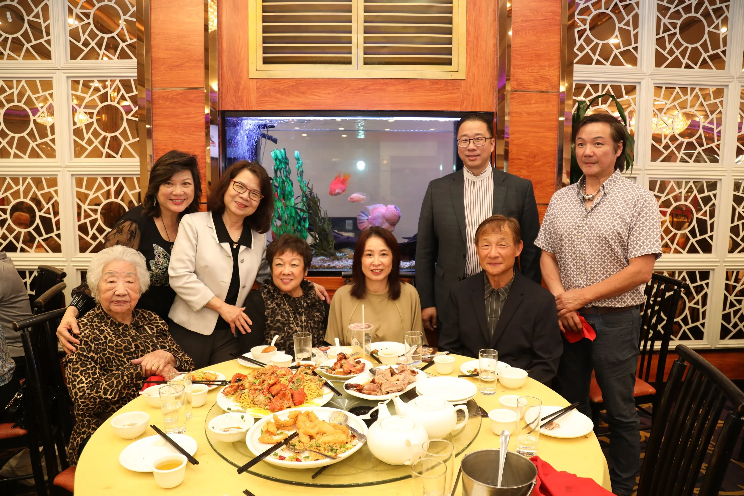 A group of seven people, three women and four men, posing around a large round table with food in a restaurant. Behind them is an aquarium with colorful fish and decorative wall panels with geometric patterns. The table has various dishes including n
