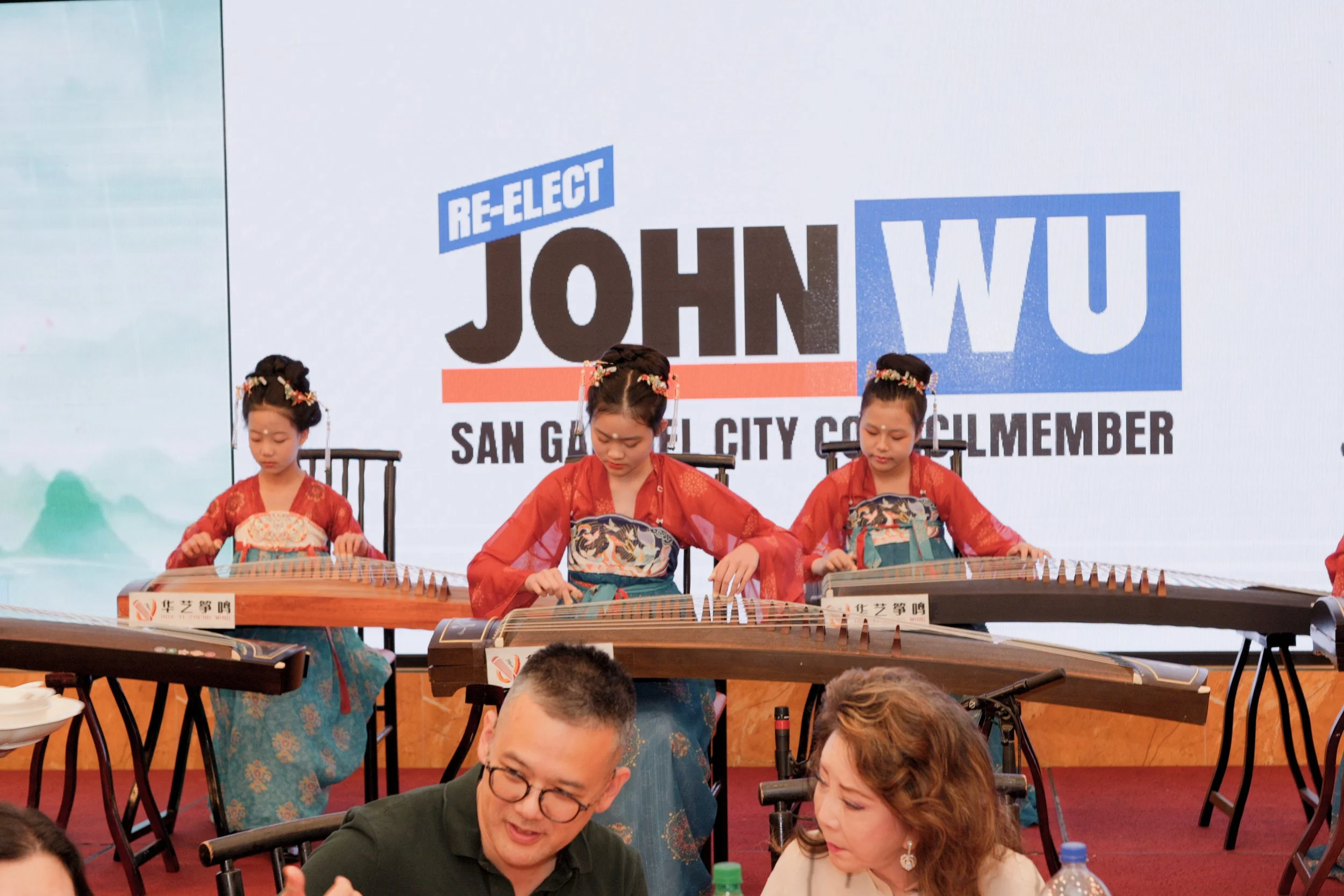 Three young girls are playing traditional Asian stringed instruments on stage, dressed in traditional red attire with floral headpieces, in front of a banner that promotes re-electing John Wu as San Gabriel City Council member.