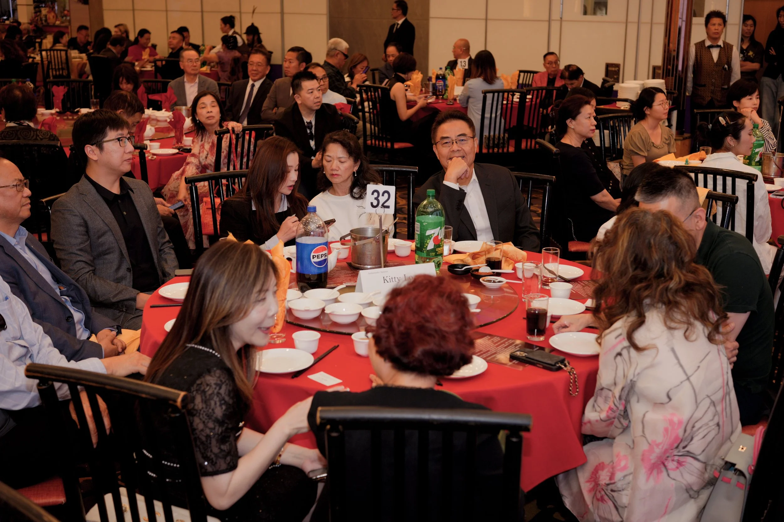 A large gathering of people seated at banquet tables in a formal setting, likely a banquet or conference, with some individuals looking at their phones and others engaged in conversation. The tables are covered with red tablecloths and are set with d