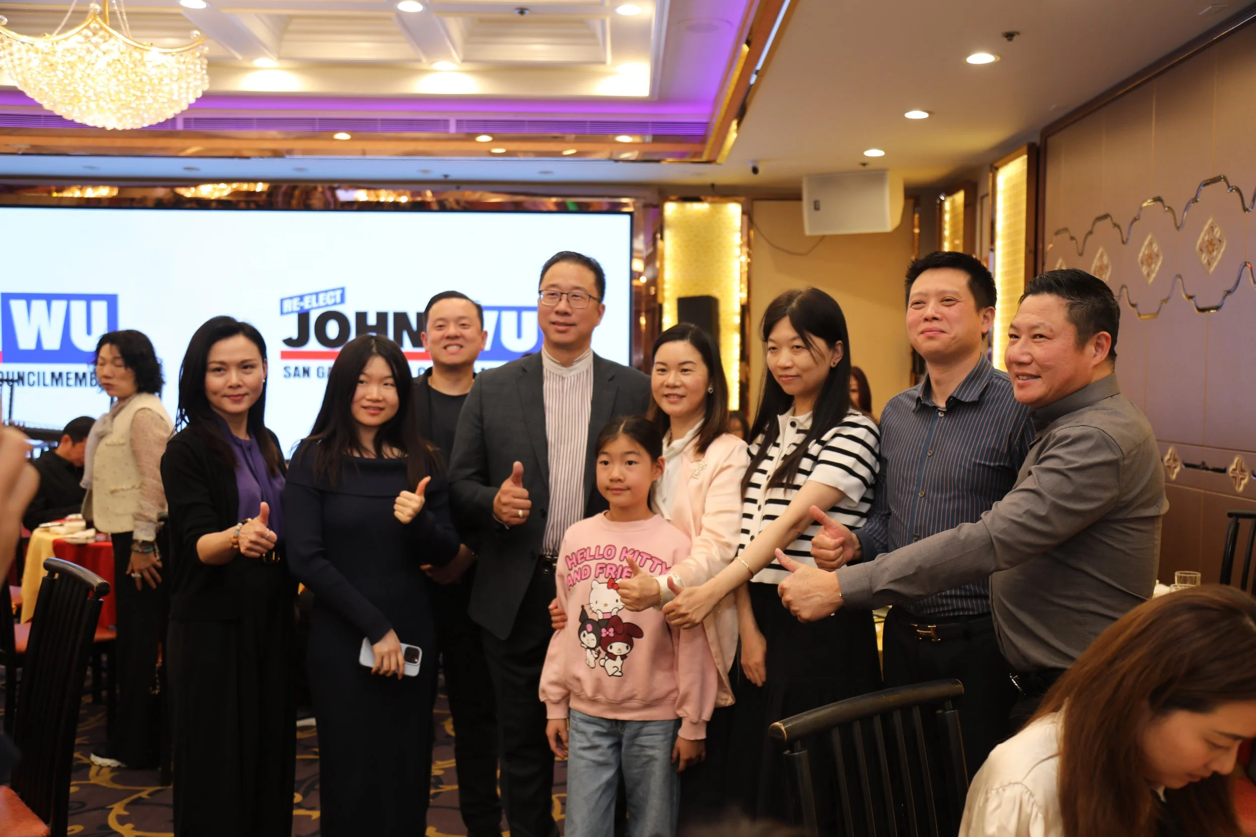 A group of people, including men, women, and children, standing together at an indoor event. In the background, a large screen displays political campaign graphics for John Yu. The setting is a banquet hall with decorative lighting and wall designs.