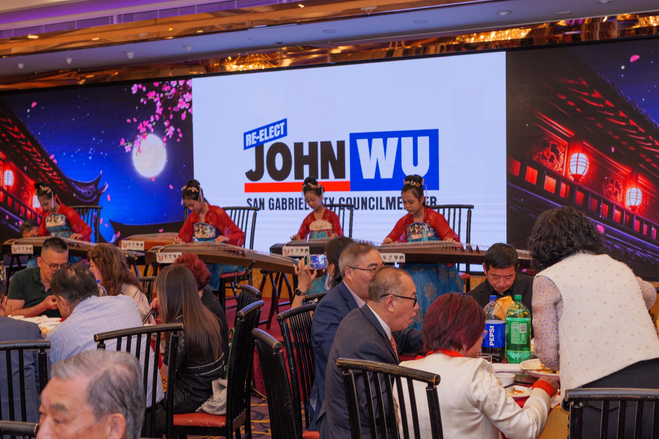 A large screen displays a campaign sign for John Wu, a candidate for San Gabrial City Council Member, with a backdrop of traditional Asian architectural and cherry blossom images. The scene shows a gathering of people, including performers in traditi
