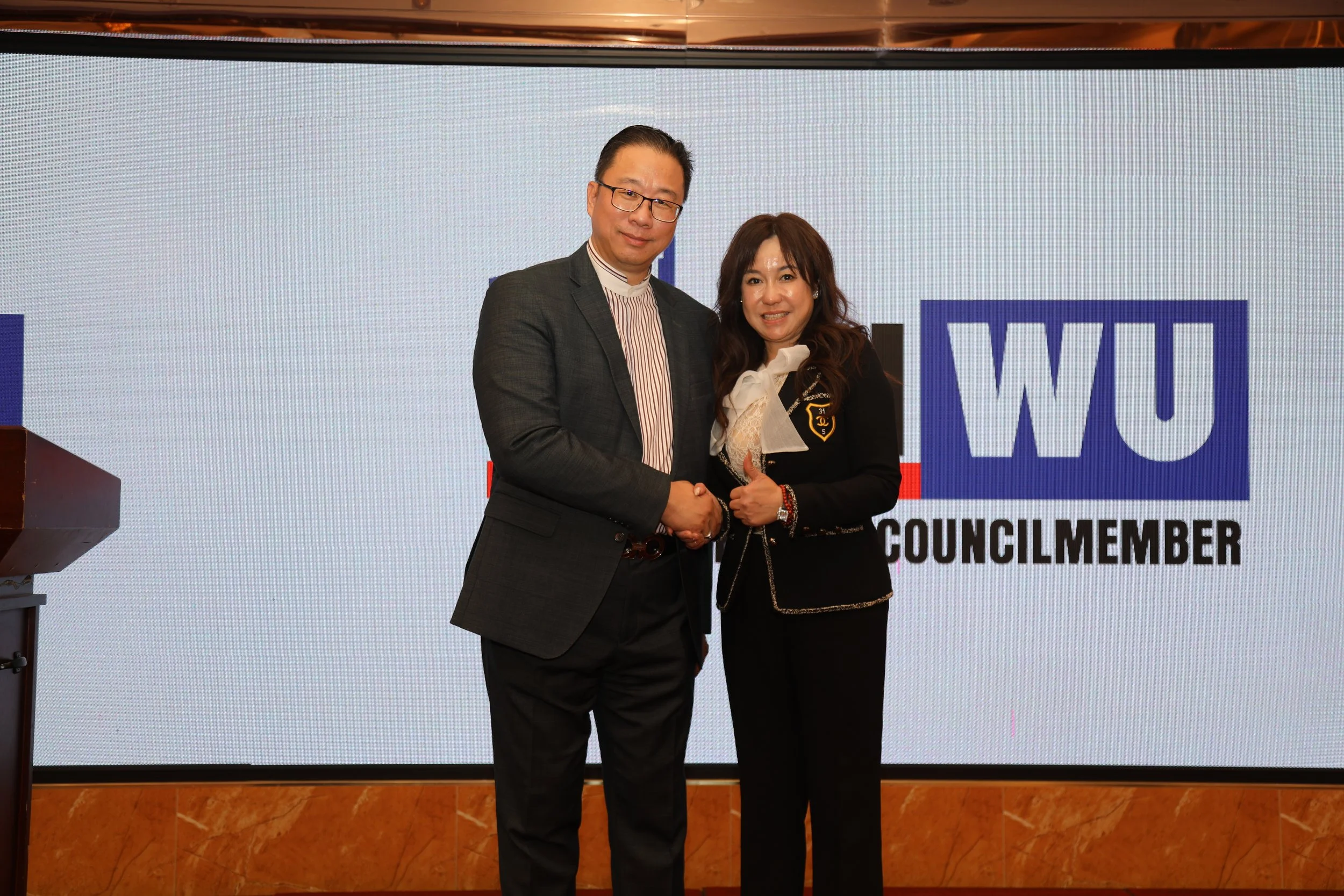 Two people, a man and a woman, shaking hands in front of a large screen that displays the text 'WUI' and 'COUNCLIMEMEBER.' The man is wearing a dark suit and glasses, and the woman is dressed in a dark blazer with a white bow tie. They are smiling.