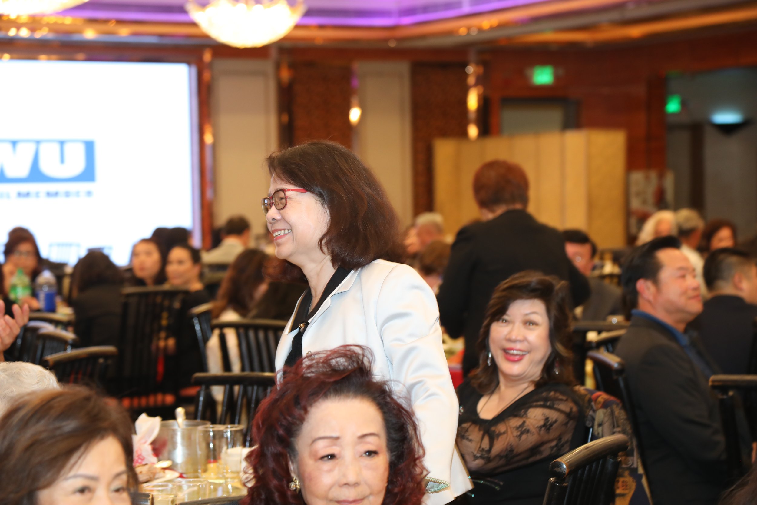 A woman with dark hair, glasses, and a white blazer smiling at an event with many people seated at tables, a large screen with a partial logo, and colorful lights on the ceiling.