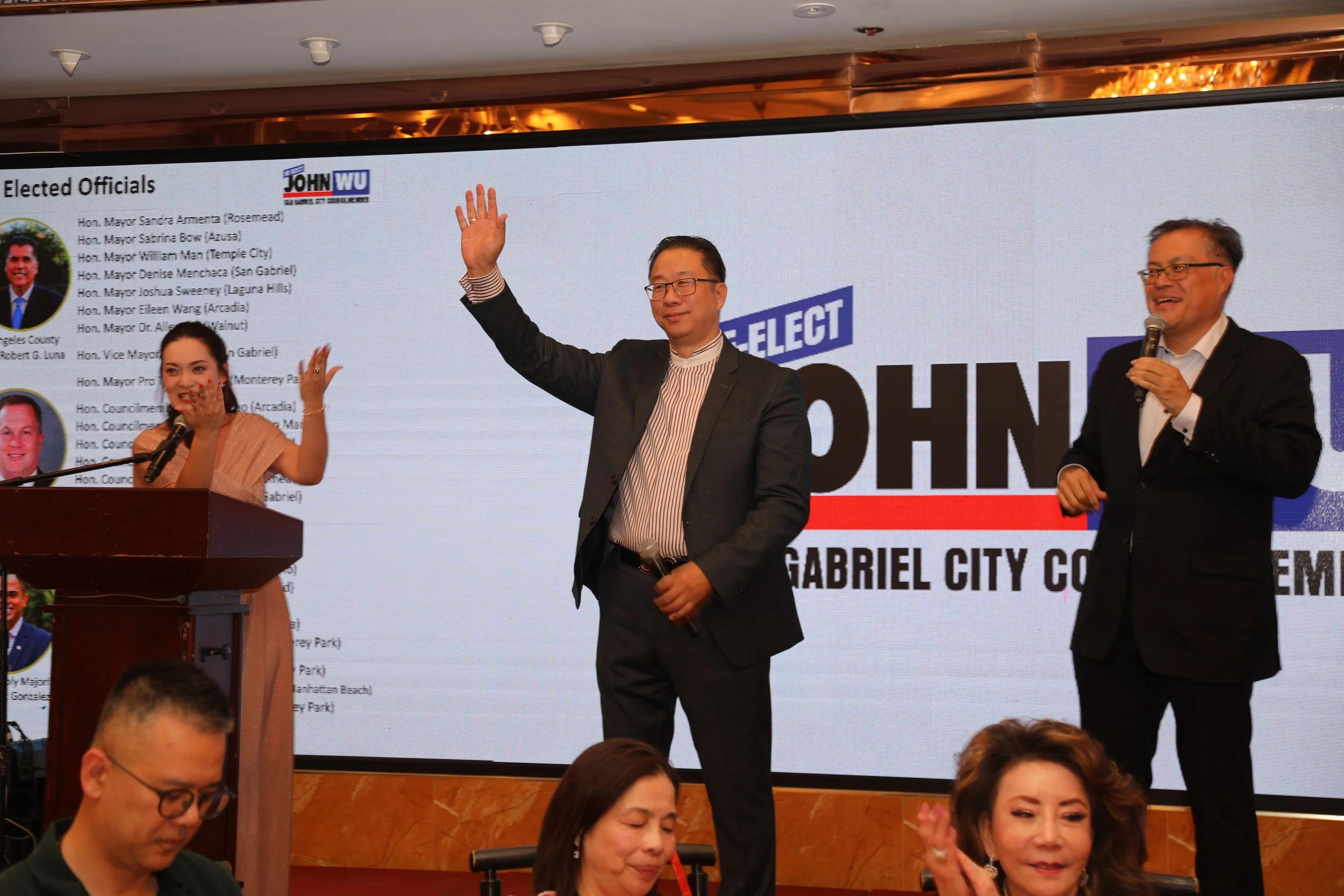 A group of politicians standing in front of a large screen at a formal event. One man is waving, another smiling while holding a microphone. Several women are seated in front, clapping and looking towards the stage.