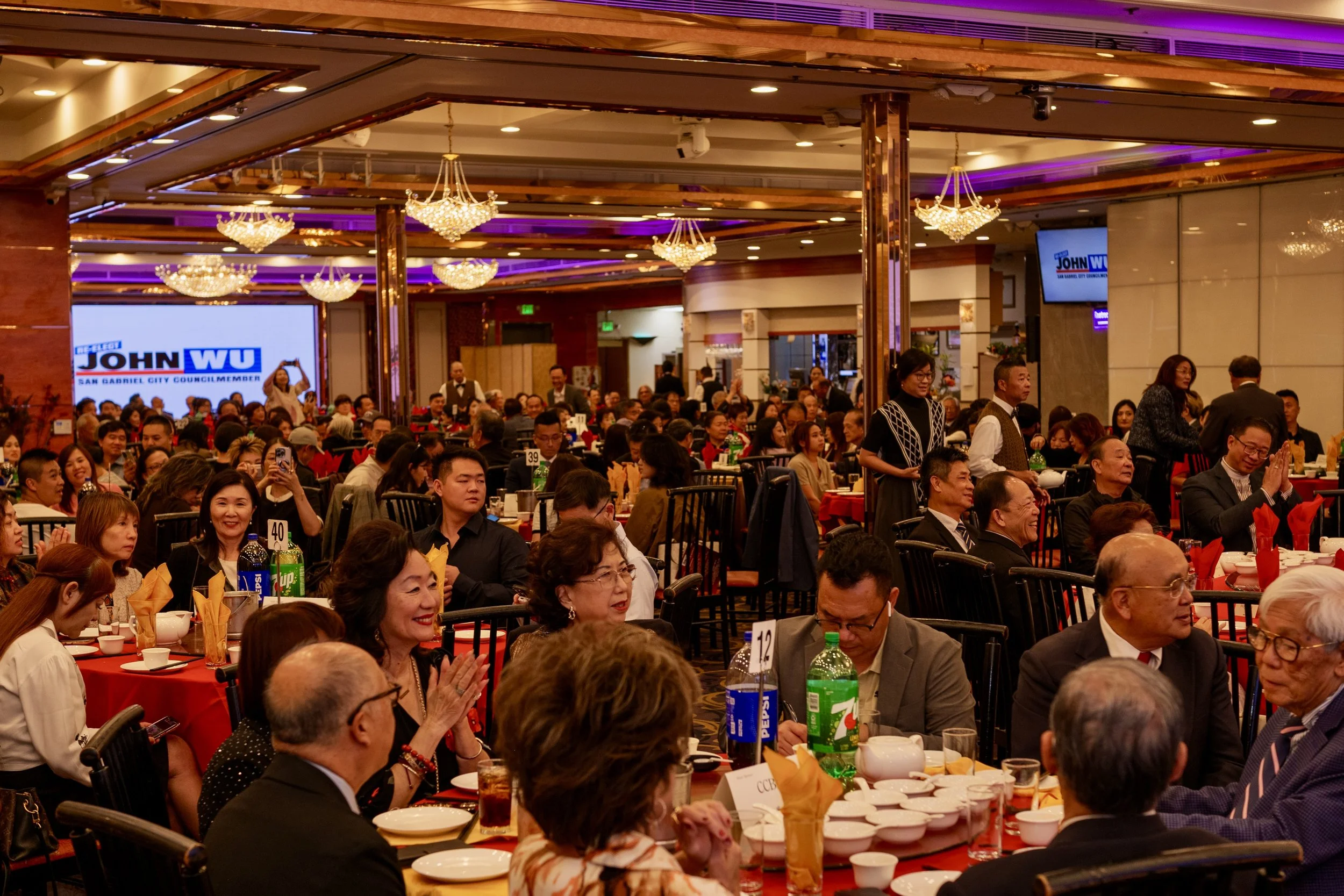 A large banquet hall filled with people seated at tables, wearing formal attire, with a screen displaying a political campaign logo in the background.