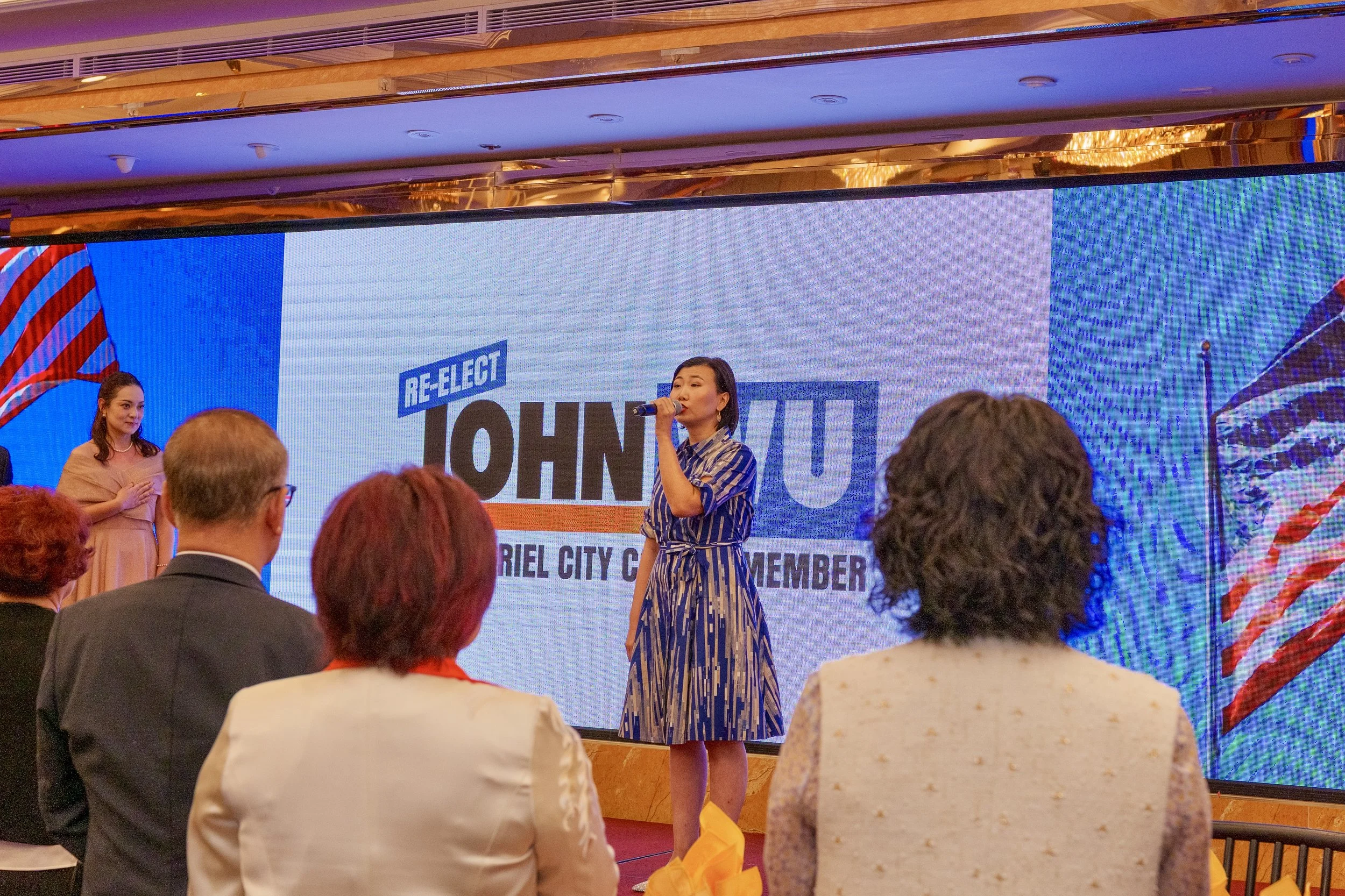 A woman speaking into a microphone on a stage with a large screen behind her that reads 'Re-elect John' and 'Griel City Council Member.' There are several people in the audience watching, including a man with gray hair and women dressed in profession