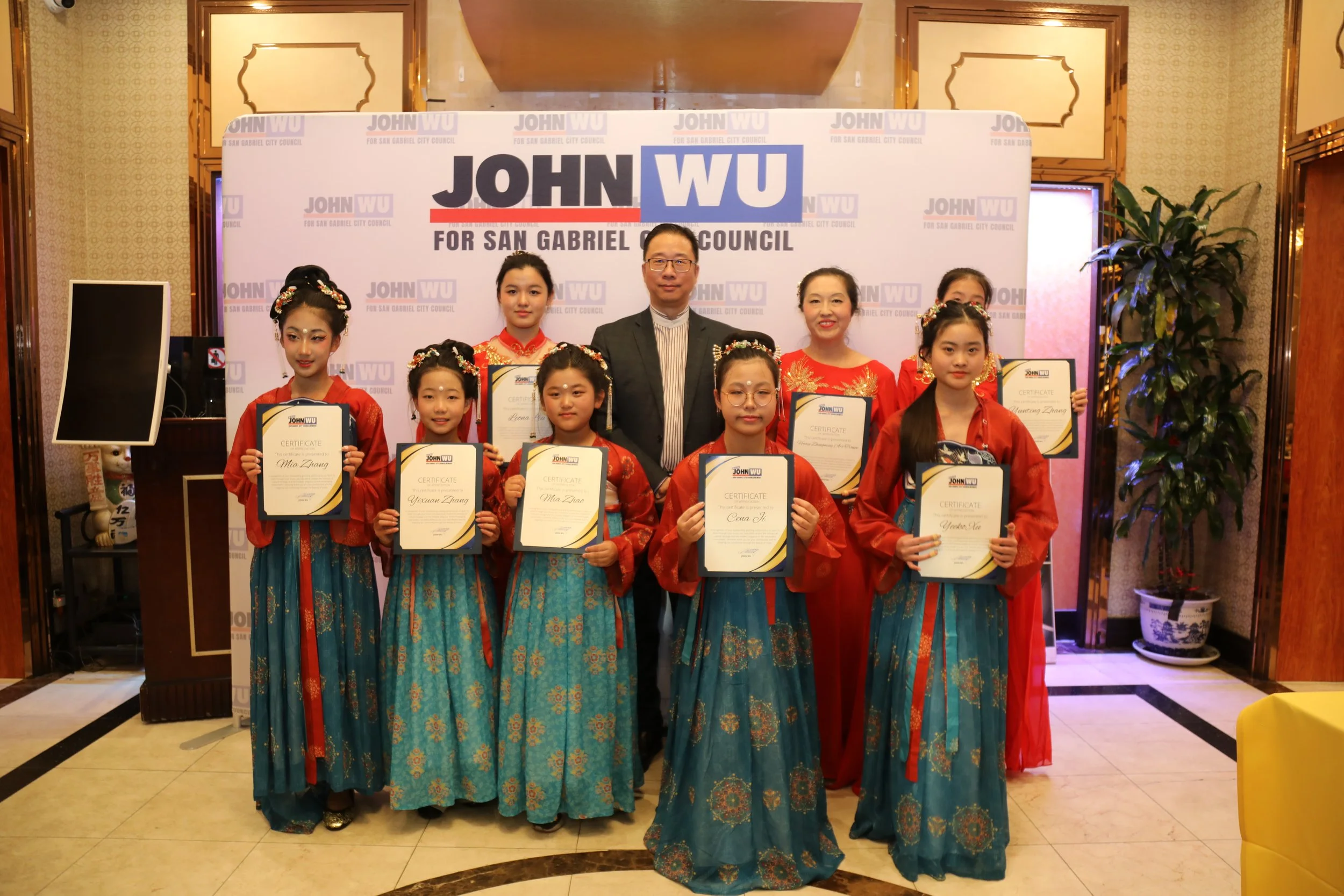 Group photo of eight women and one man standing in front of a backdrop with campaign logos. The women are dressed in traditional Chinese attire and holding certificates. The setting appears to be an indoor event space decorated with plants and a patt