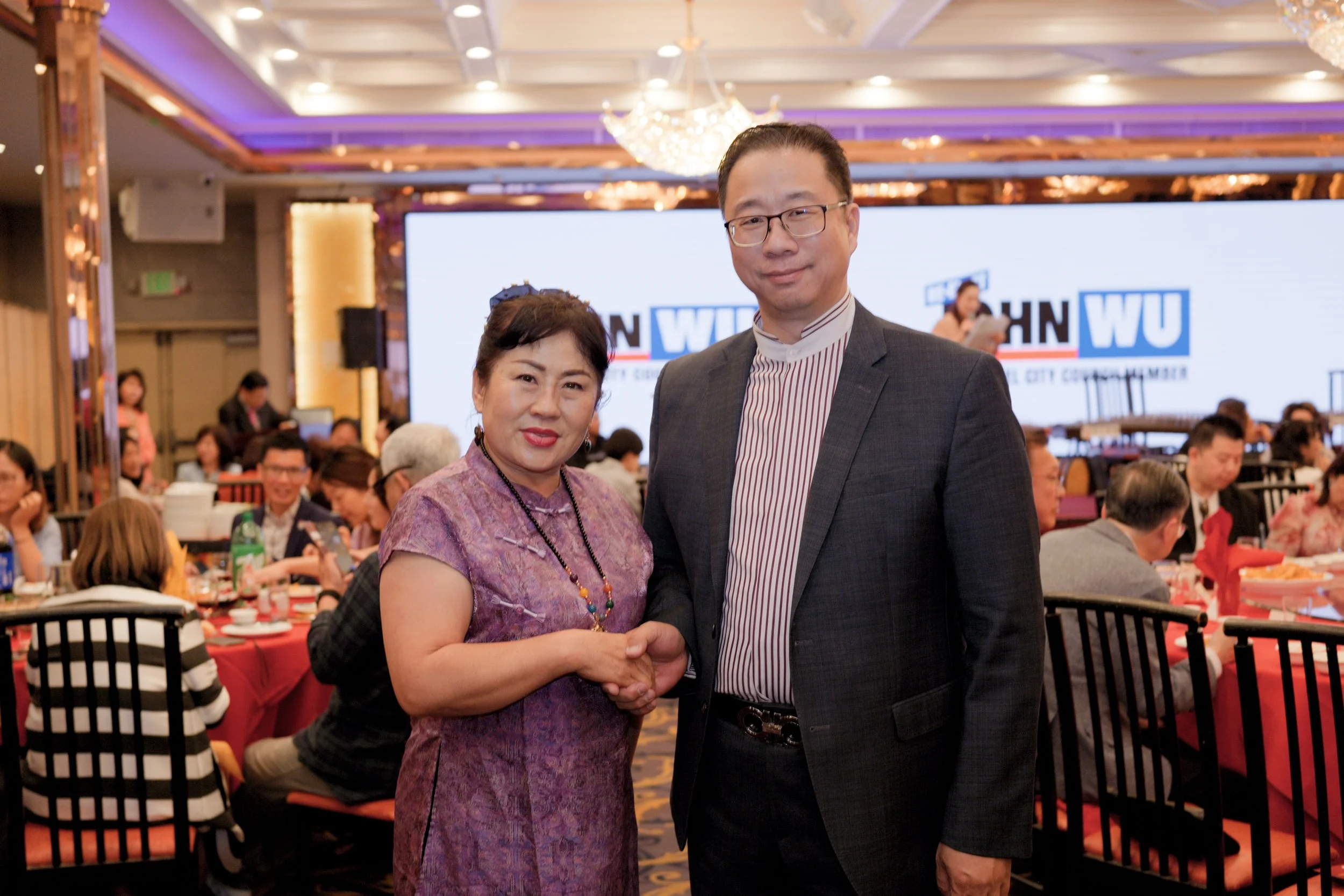 Two people shaking hands at a formal event server in a decorated banquet hall filled with seated attendees and a large screen displaying the WU logo.