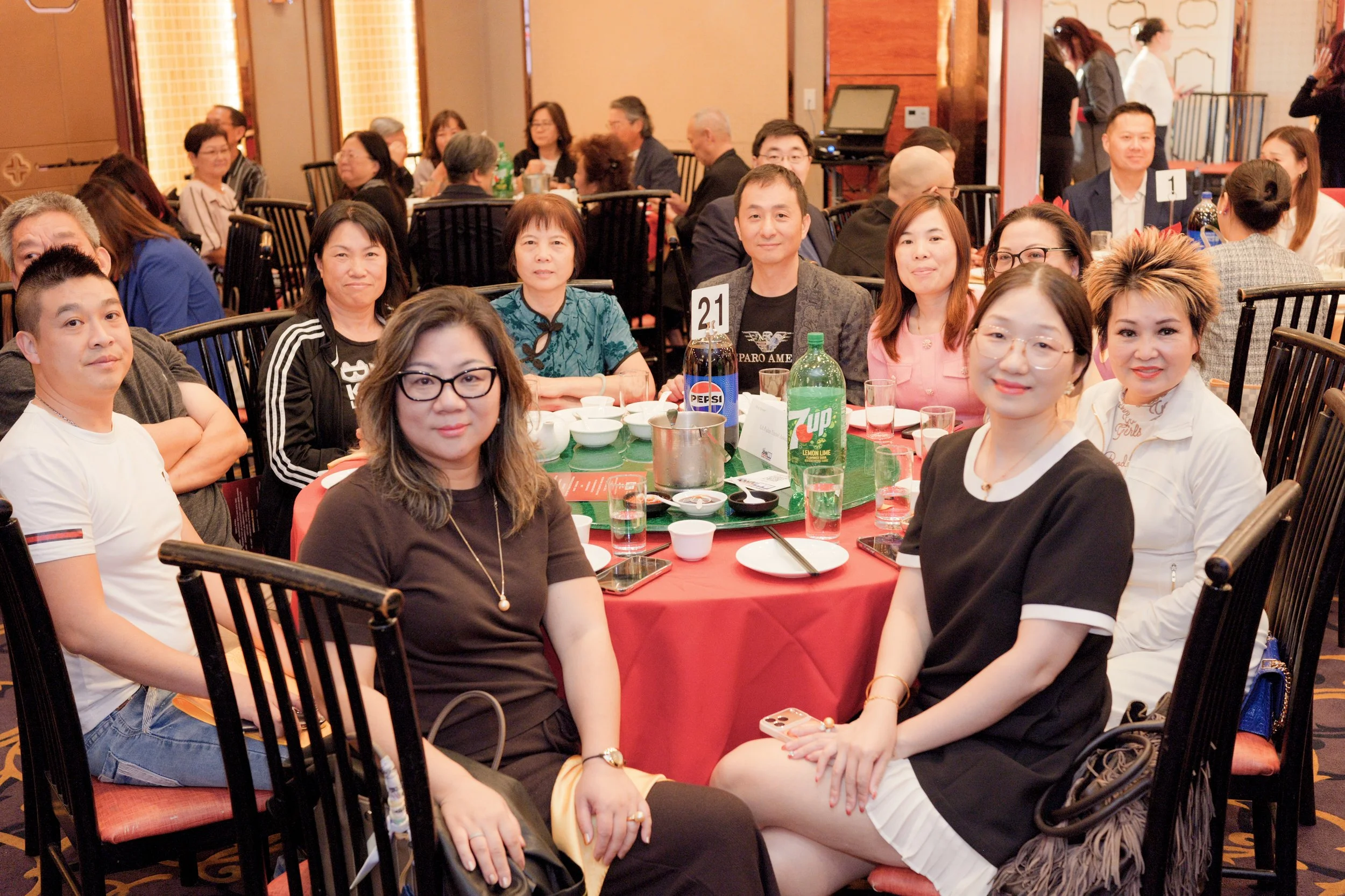A diverse group of people sitting around a round table at a banquet or conference room, smiling for a photo. The table has drinks and food, with a sign indicating table 21.