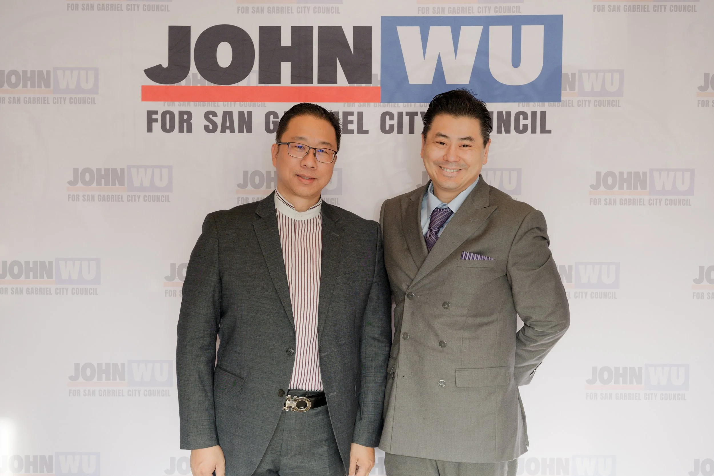 Two men dressed in business suits standing in front of a backdrop that reads "John WU for San Gabriel City Council." The man on the left has glasses, short dark hair, and is wearing a dark plaid suit with a striped shirt. The man on the right has dar