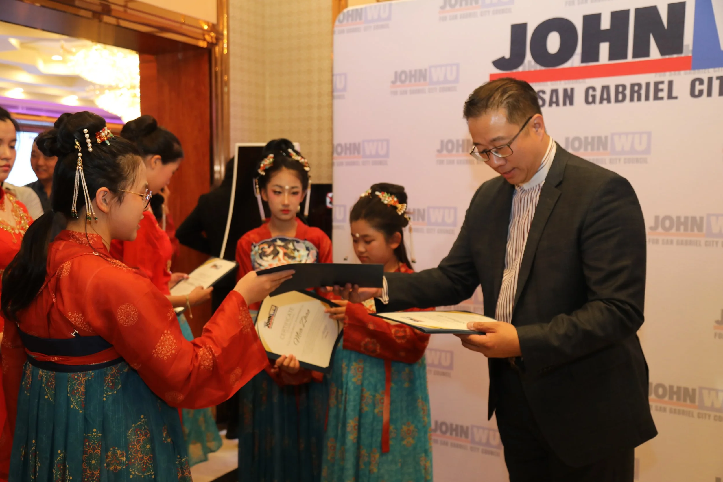 A man in a suit receiving an award from a woman in traditional Asian attire with others in traditional clothing standing nearby during an awards ceremony.