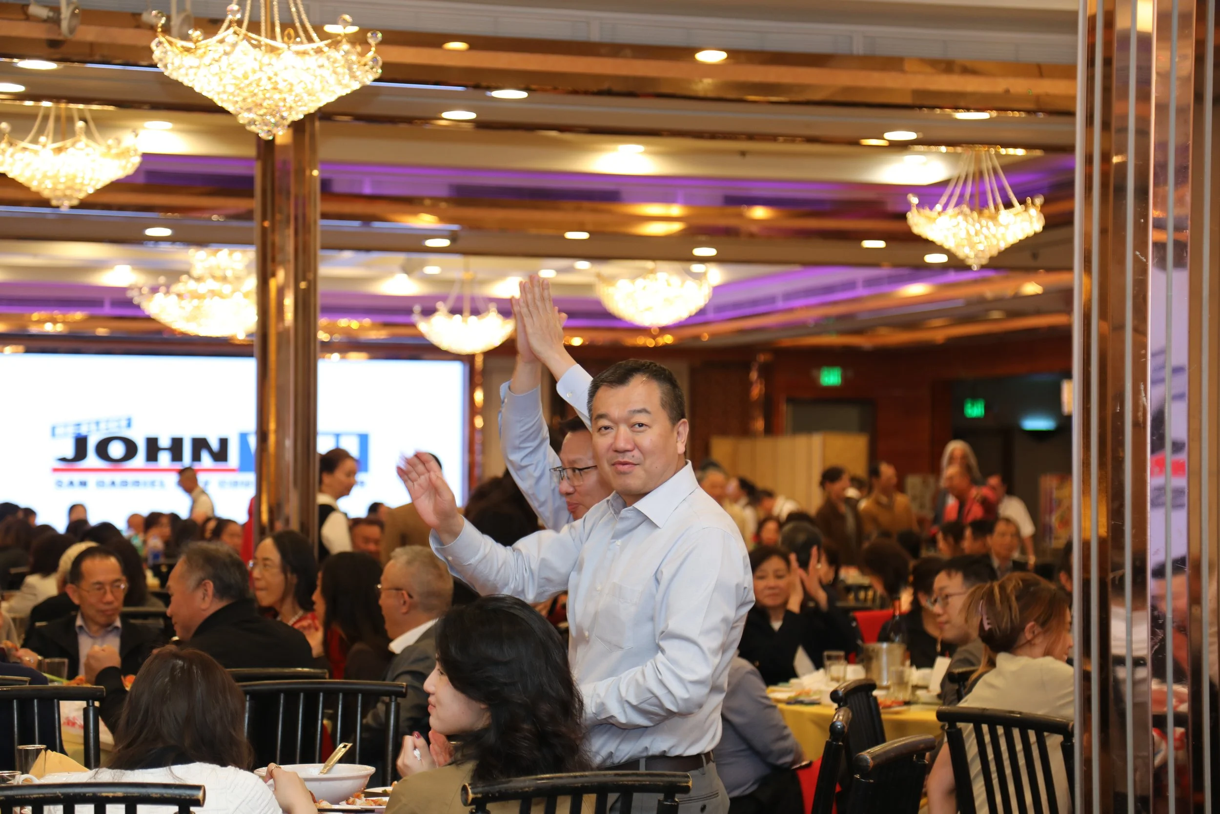 A man in a white shirt waving at a crowded banquet hall filled with people sitting at tables. The hall has wooden beams and chandeliers hanging from the ceiling.