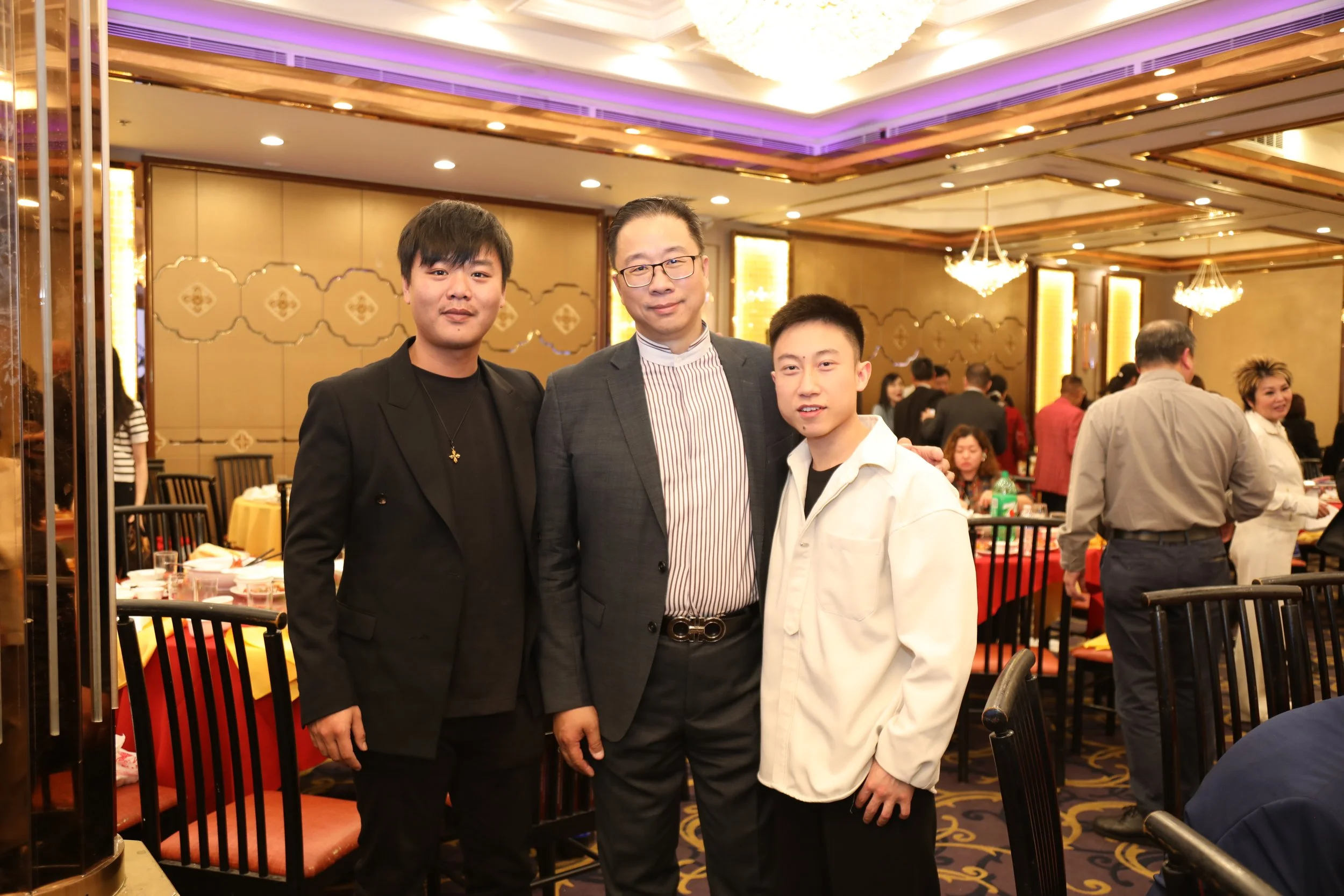 Three men standing together in a banquet hall, smiling at the camera. The hall has gold and purple decor, chandeliers, and dining tables with red tablecloths and chairs.