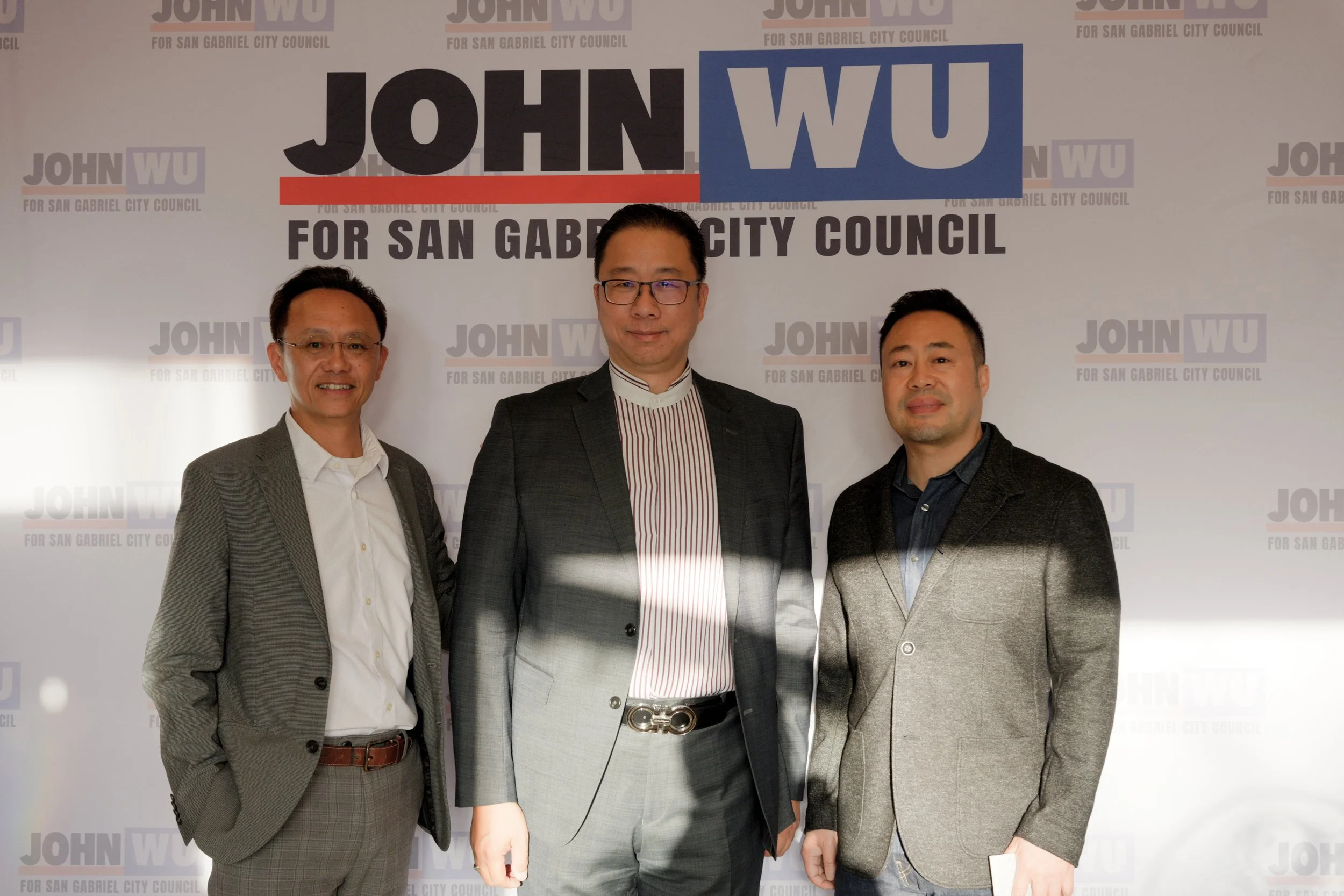 Three men dressed in business attire standing in front of a backdrop with the words 'JOHN WU FOR SAN GABRIEL CITY COUNCIL' written repeatedly.